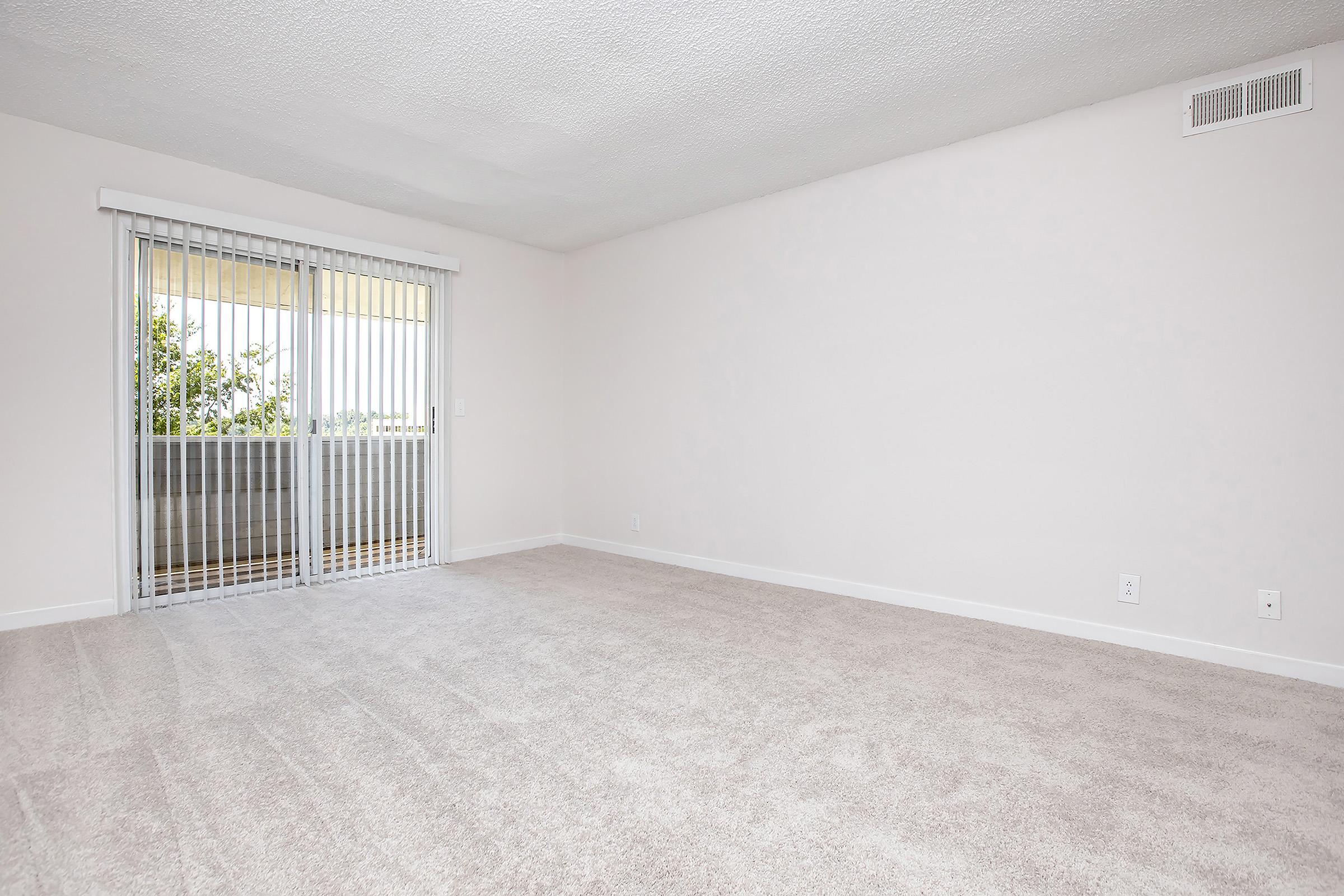 A spacious, empty room with light beige carpet and plain white walls. Large sliding glass doors lead to a small balcony, allowing natural light to flow in. The area is clean and uncluttered, creating a bright and airy atmosphere.