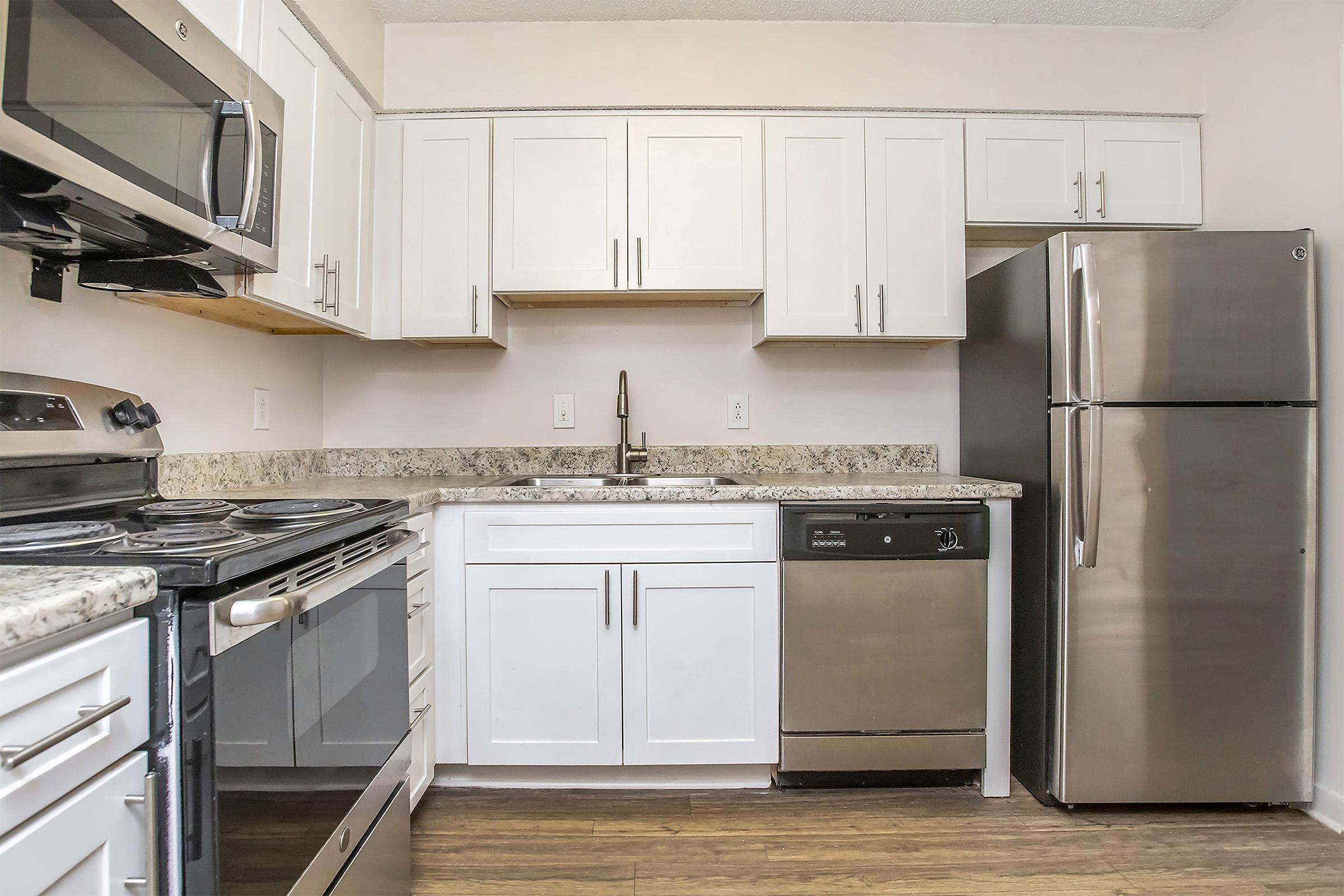 Modern kitchen featuring white cabinetry, stainless steel appliances including a microwave, oven, dishwasher, and refrigerator. The countertop is made of granite, and the space has a light, airy feel with a wooden floor. The layout is efficient with ample storage and a sleek design.