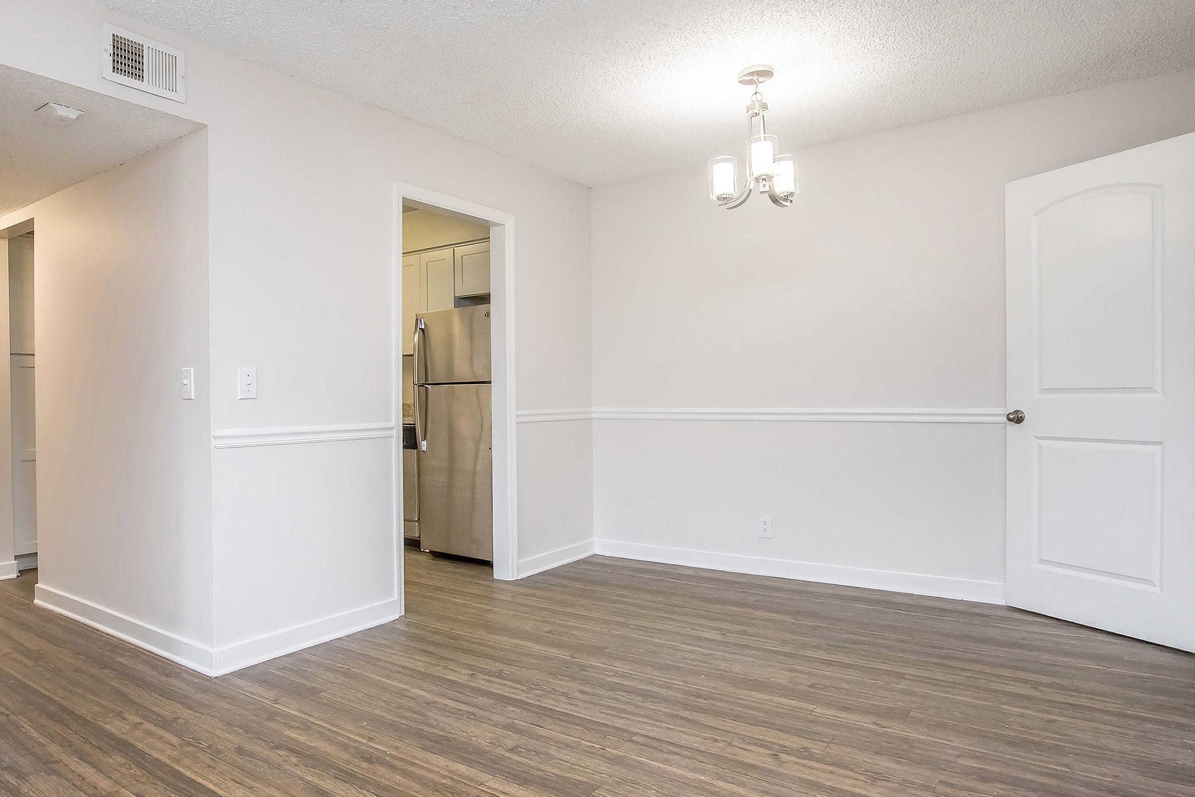 A well-lit, empty room featuring light-colored walls and wooden flooring. To the left, a doorway leads to a kitchen with stainless steel appliances visible. A simple chandelier hangs from the ceiling, and there is a door on the right, possibly leading to another room. The space is clean and modern, ready for furniture.