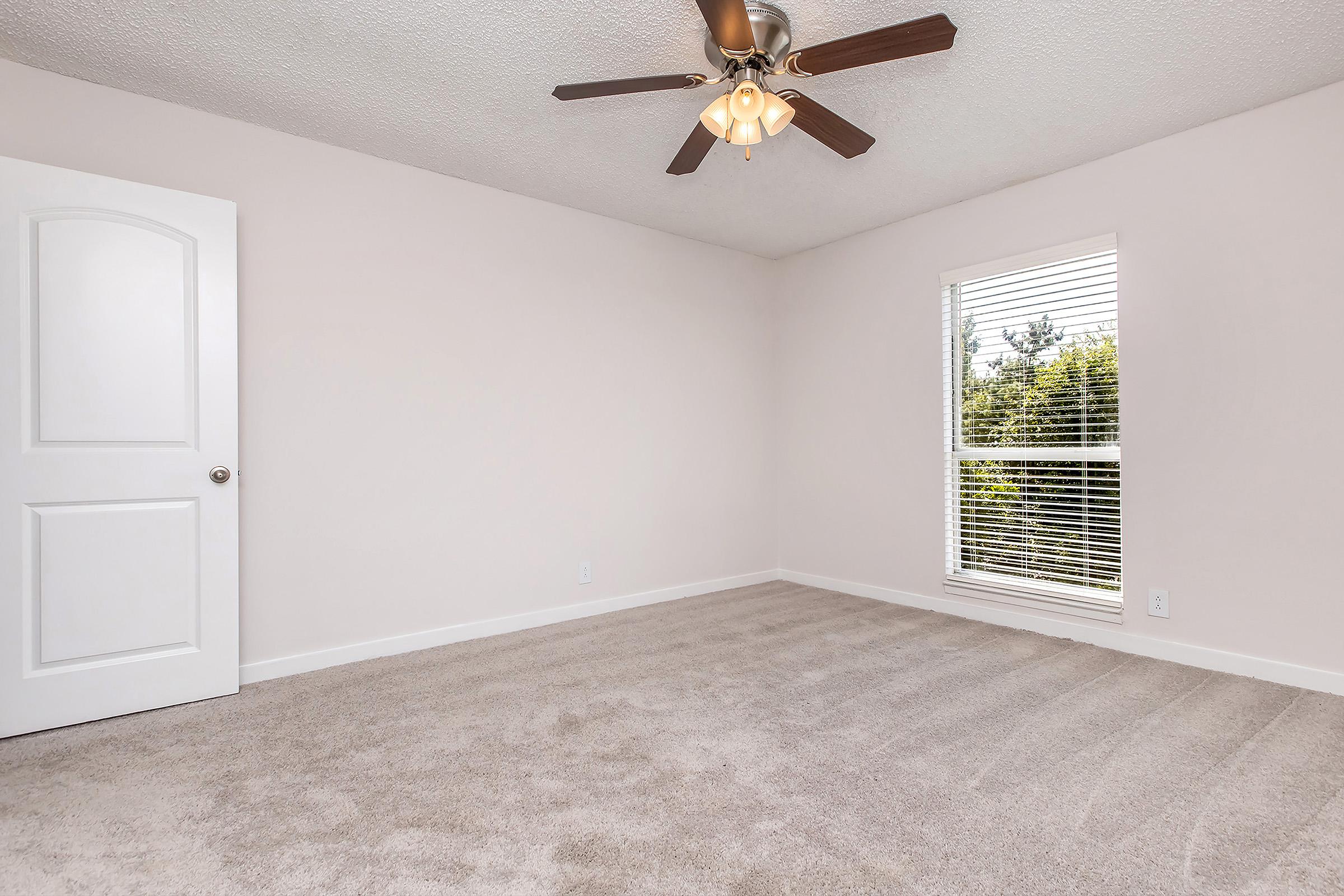 A light-colored bedroom featuring beige carpet, a ceiling fan with a light fixture, and a white door. There is a window with blinds allowing natural light to enter, revealing a view of greenery outside. The walls are painted a soft shade, creating a bright and inviting atmosphere.