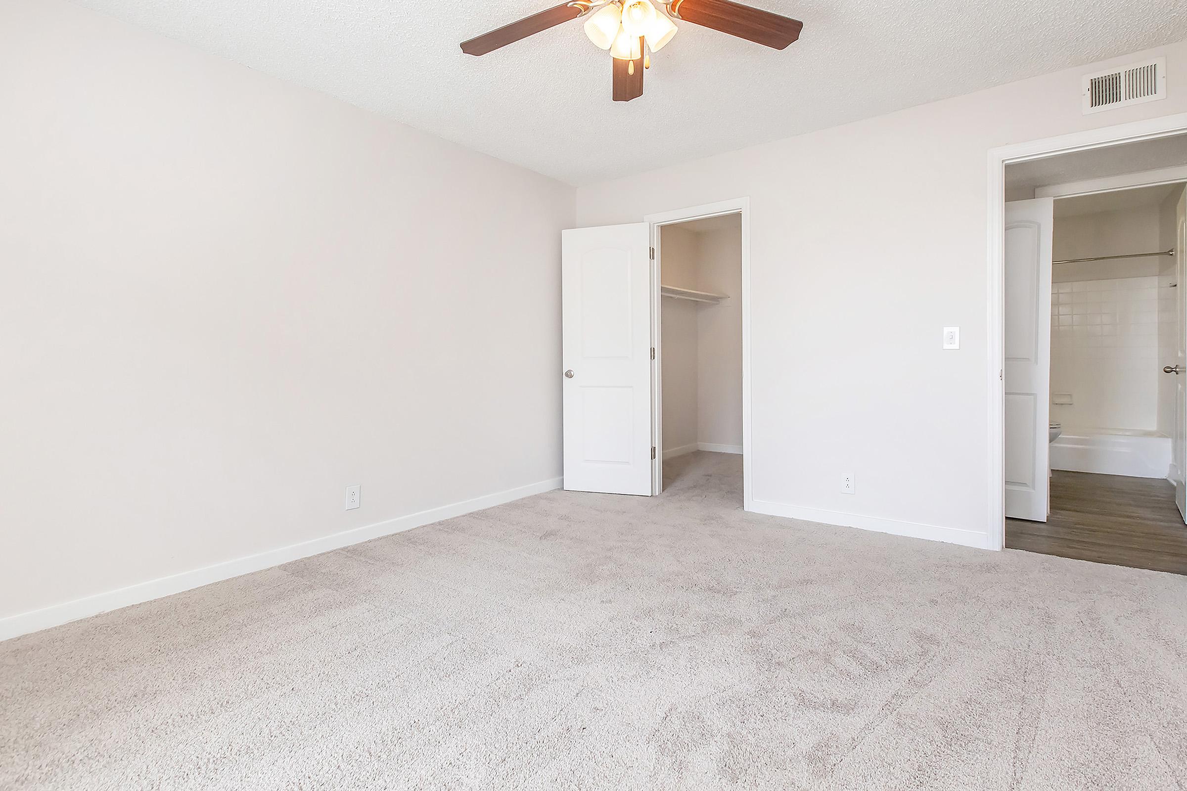 A spacious, empty bedroom featuring light-colored walls and beige carpet. A light ceiling fan hangs from the ceiling, and there is a closet with an open door to the left. A bathroom can be glimpsed through a second door on the right, adding to the room's overall bright and inviting atmosphere.