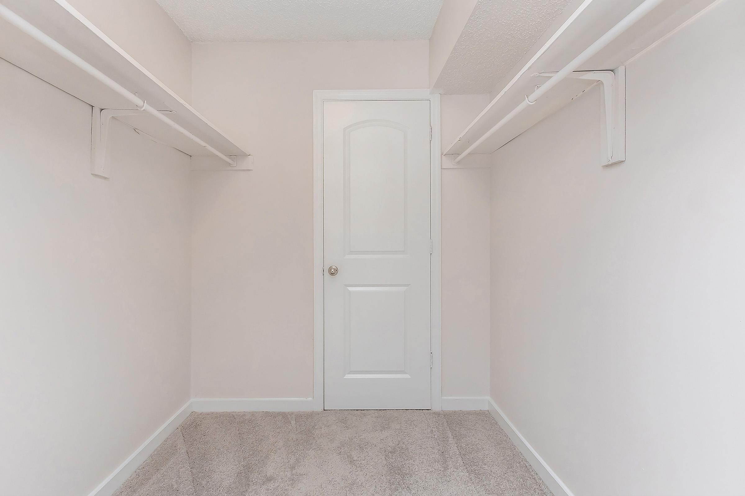 Empty closet with beige walls and light carpet. Two hanging rods are positioned on the left and right sides, and a closed white door is visible at the back. The space is well-lit and appears spacious, suitable for storage or organization.