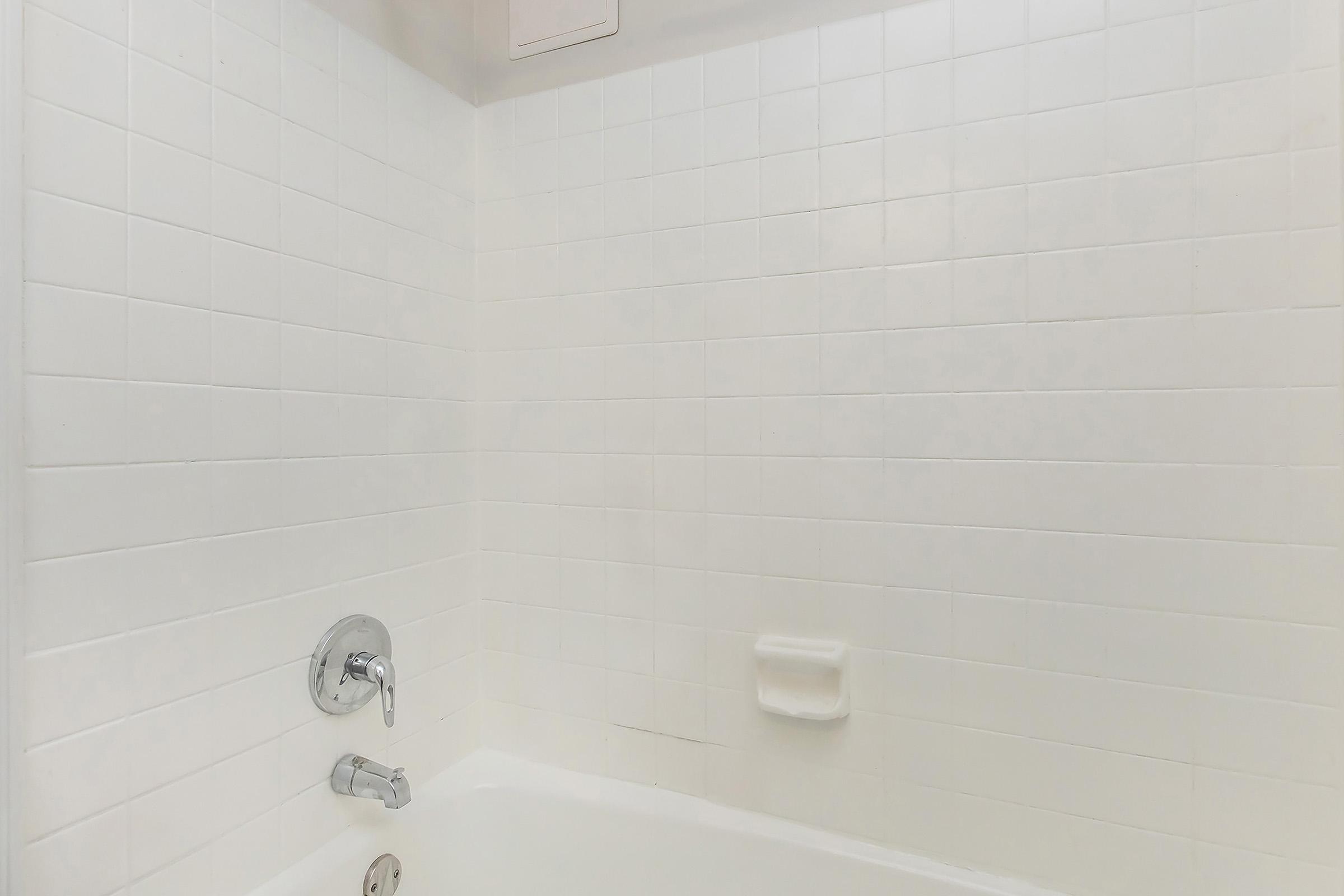 A clean, white tiled bathroom wall with a bathtub visible at the bottom. The wall features a faucet and a detachable showerhead, while a soap dish is mounted on the wall. The overall appearance is bright and minimalistic.