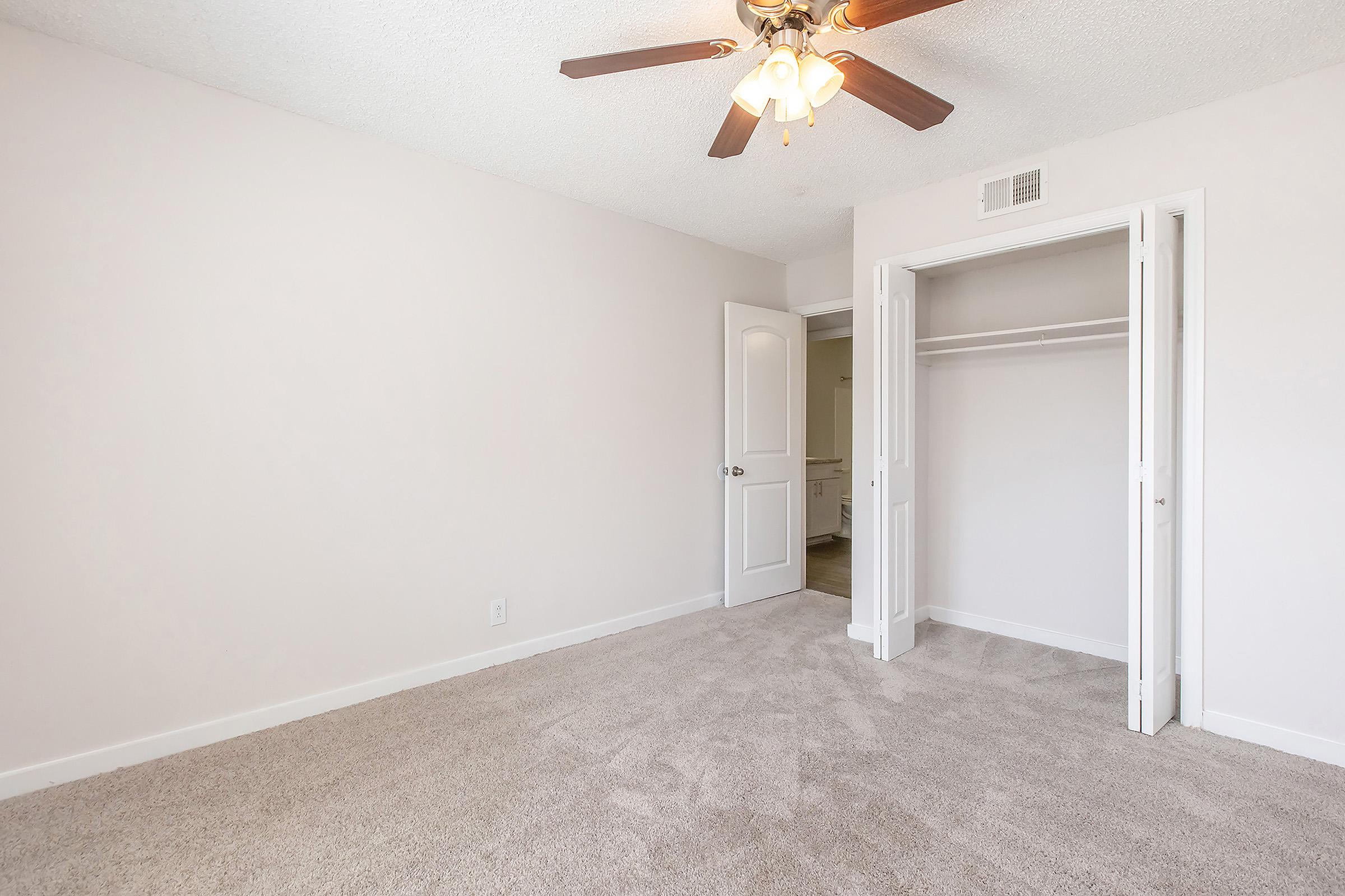A bright and spacious empty bedroom featuring light-colored walls, a ceiling fan with light fixture, and soft carpet. There is a closed door leading to an adjoining room and a closet with sliding doors on one side. The overall atmosphere is clean and inviting, ideal for personal customization.