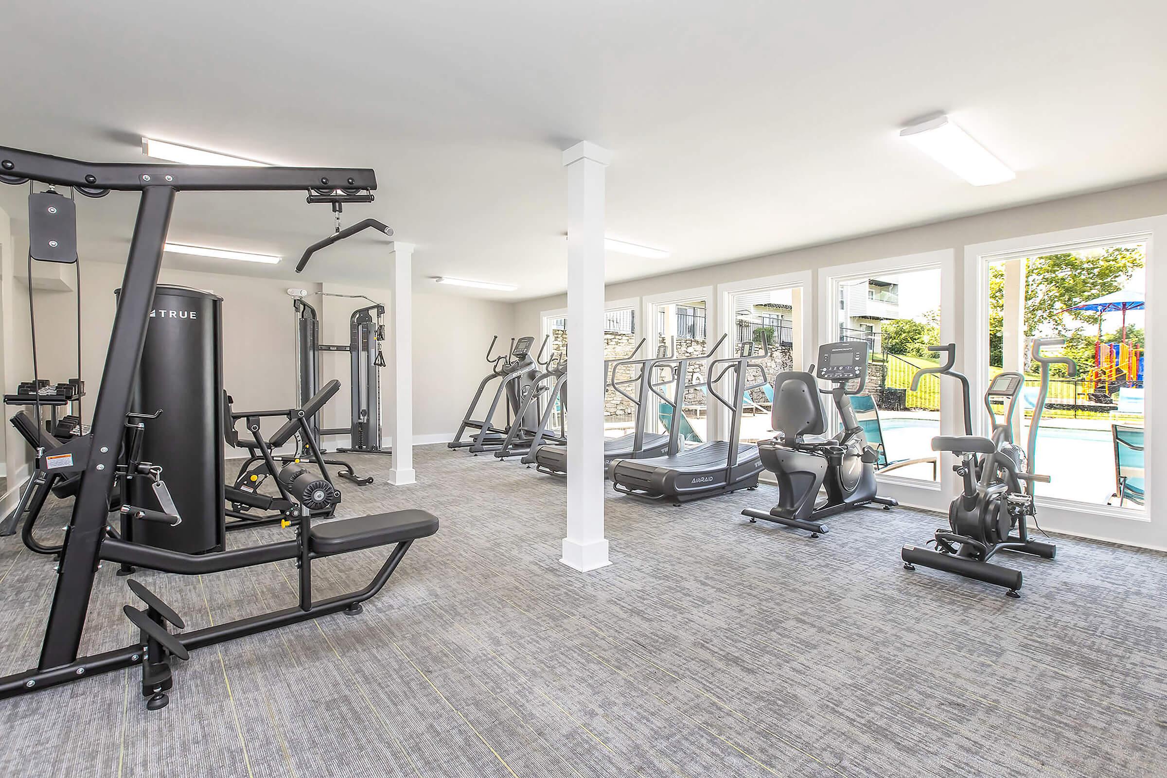 A modern fitness room featuring various exercise equipment, including treadmills, stationary bikes, strength training machines, and free weights. The space is well-lit with large windows offering a view of an outdoor area. The flooring is carpeted, creating a comfortable workout environment.