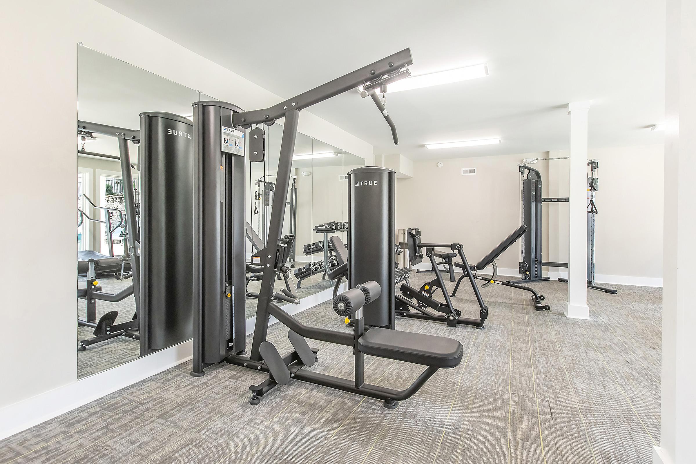 A modern gym interior featuring various workout equipment, including a multi-station weight machine, free weights, and exercise benches. The bright space has large mirrors and neutral-colored walls, creating a clean and inviting atmosphere for fitness activities.