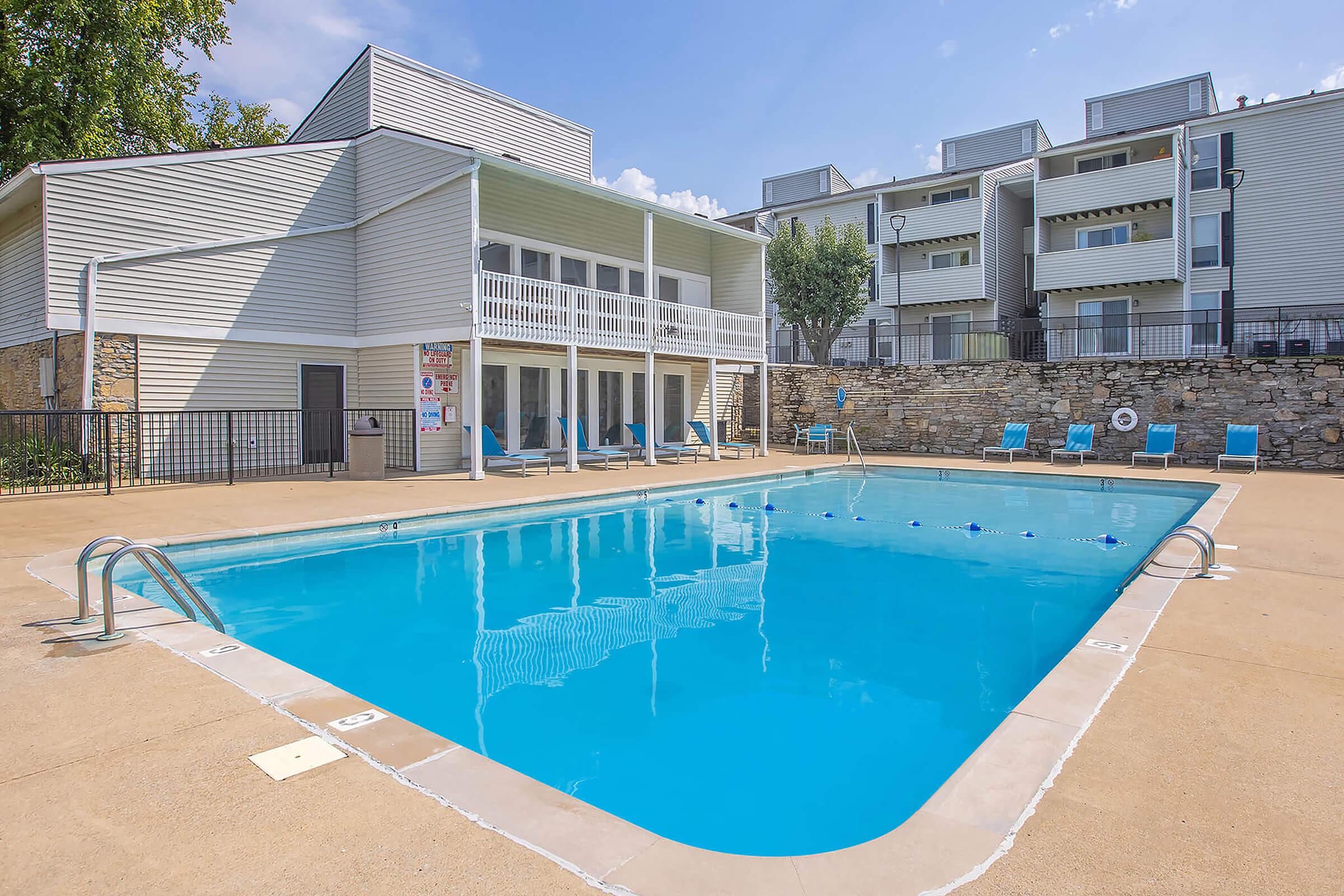 A clear swimming pool surrounded by a concrete deck, with lounge chairs positioned around it. In the background, a two-story building with a balcony and several apartments is visible, along with trees and a stone wall enclosing the area. The sky is bright and sunny.
