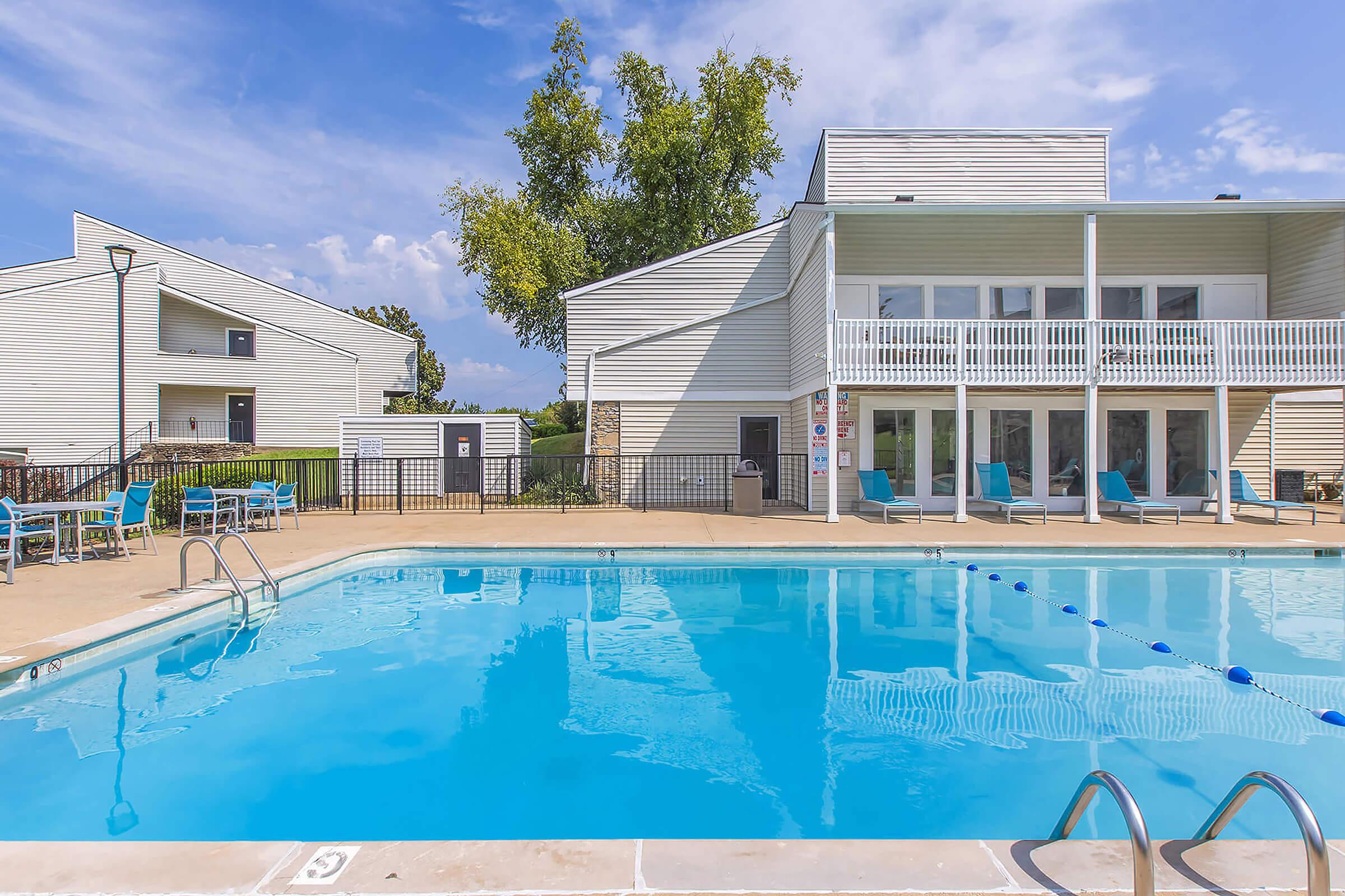 A clear swimming pool surrounded by a deck with lounge chairs, situated in a residential area. White buildings with balconies are visible in the background under a partly cloudy sky, creating a relaxing atmosphere.