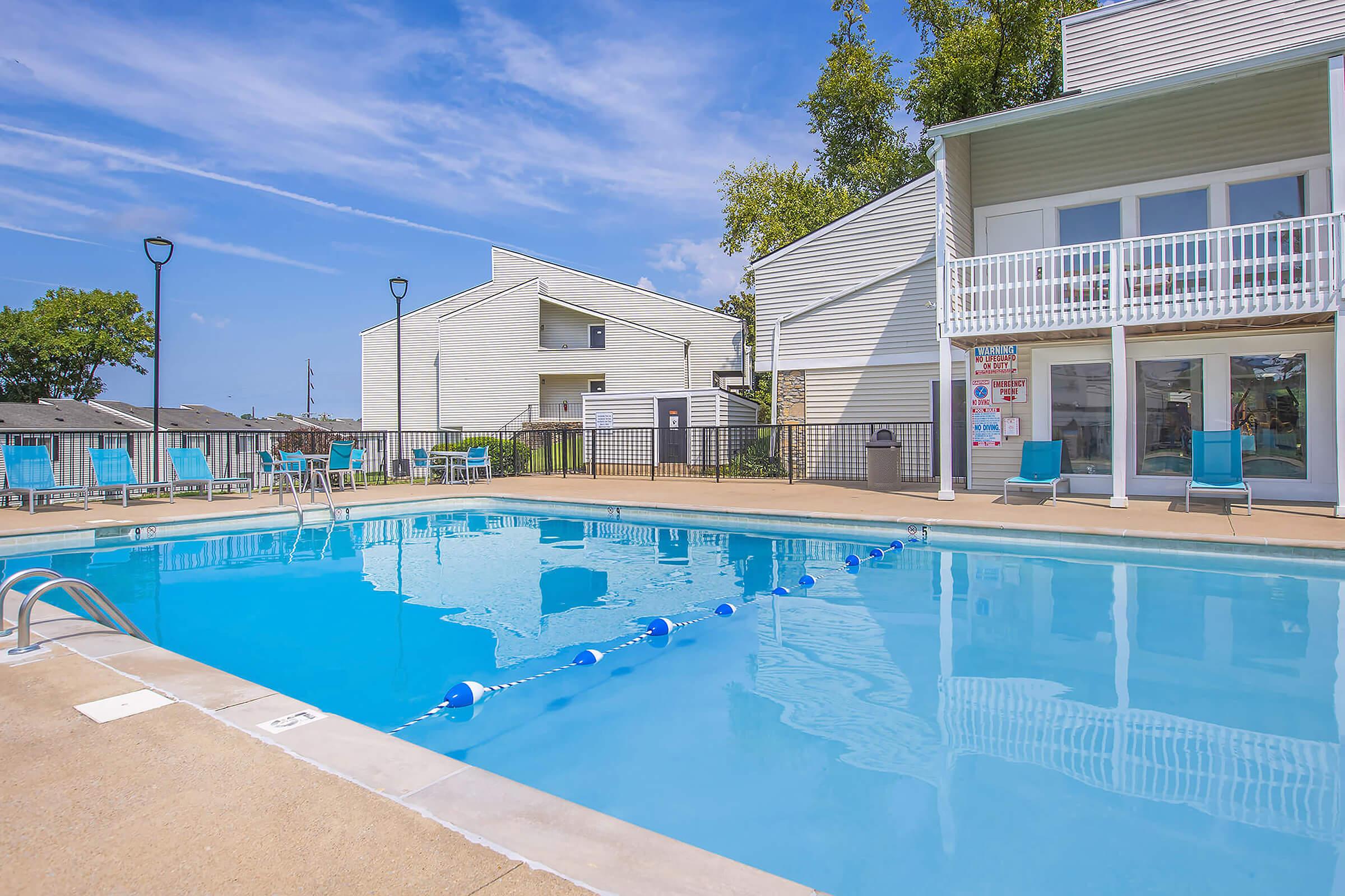 A residential outdoor swimming pool with clear blue water, lined with lounge chairs under a bright blue sky. Nearby buildings feature light-colored siding, and a fence encloses the pool area. A few trees provide shade, and the scene suggests a relaxing atmosphere.