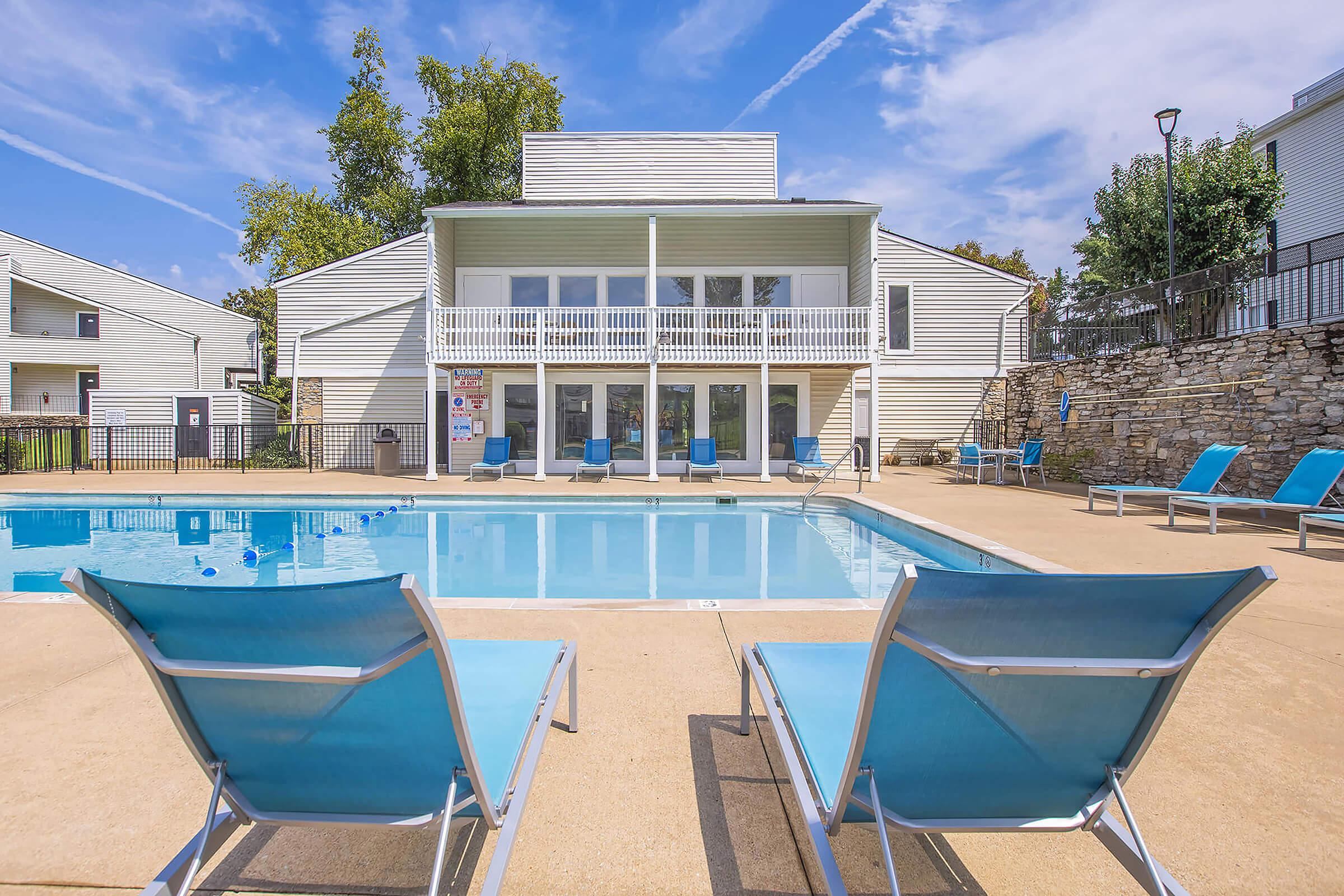 A clear swimming pool surrounded by lounge chairs in a residential area, with a two-story building in the background featuring large windows and a balcony. The sky is blue with scattered clouds, and trees are visible on the property.