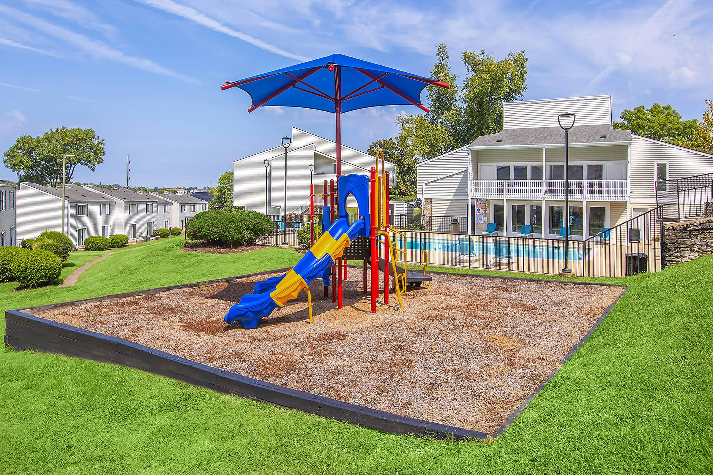 A colorful playground featuring a slide and climbing structure under a blue umbrella, set on a gravel surface. In the background, there are two-story apartment buildings and a swimming pool surrounded by grassy areas on a sunny day.