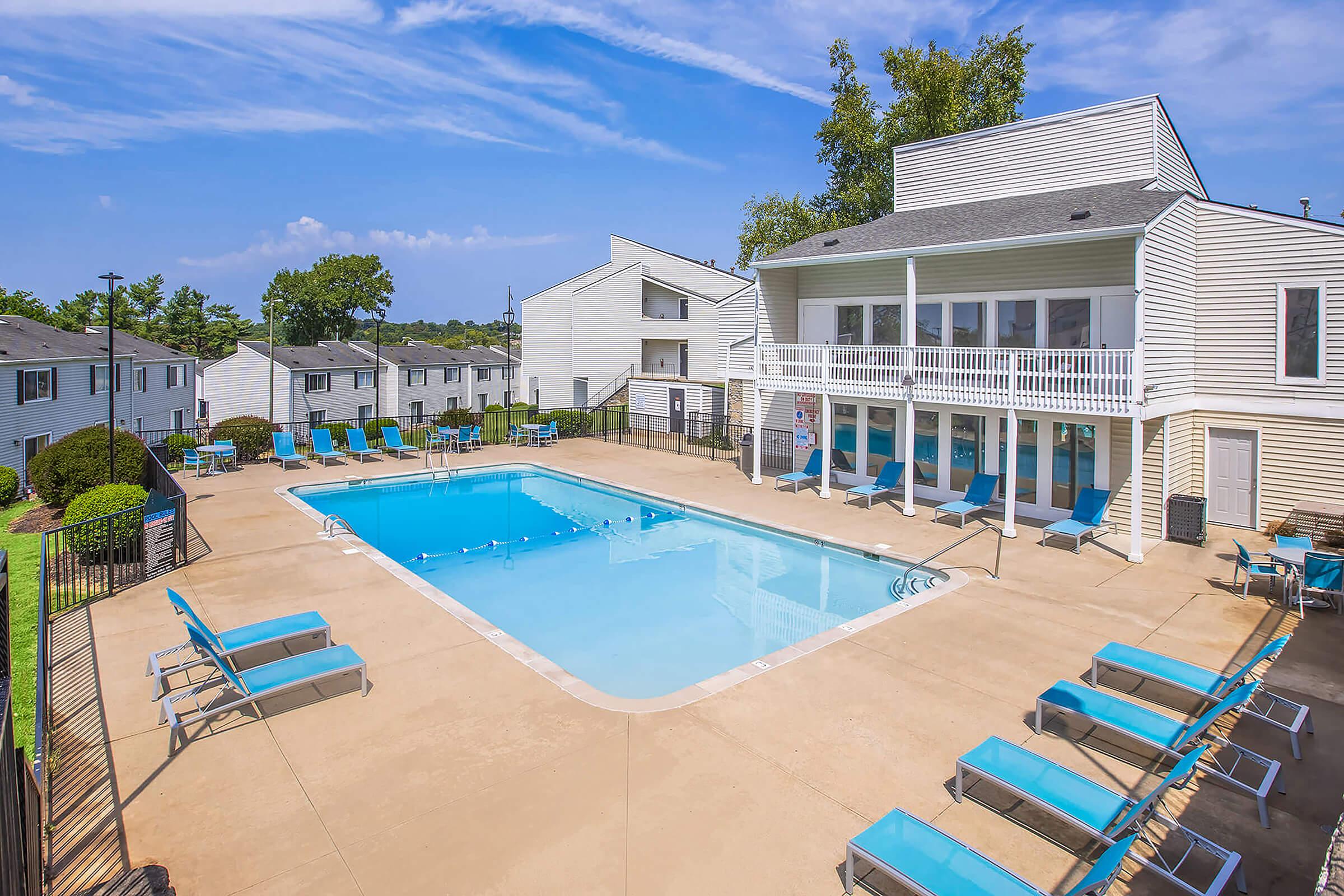 A bright, sunny view of a swimming pool surrounded by lounge chairs, with a multi-story building in the background. The scene features green trees and a clear blue sky, creating a relaxing atmosphere.