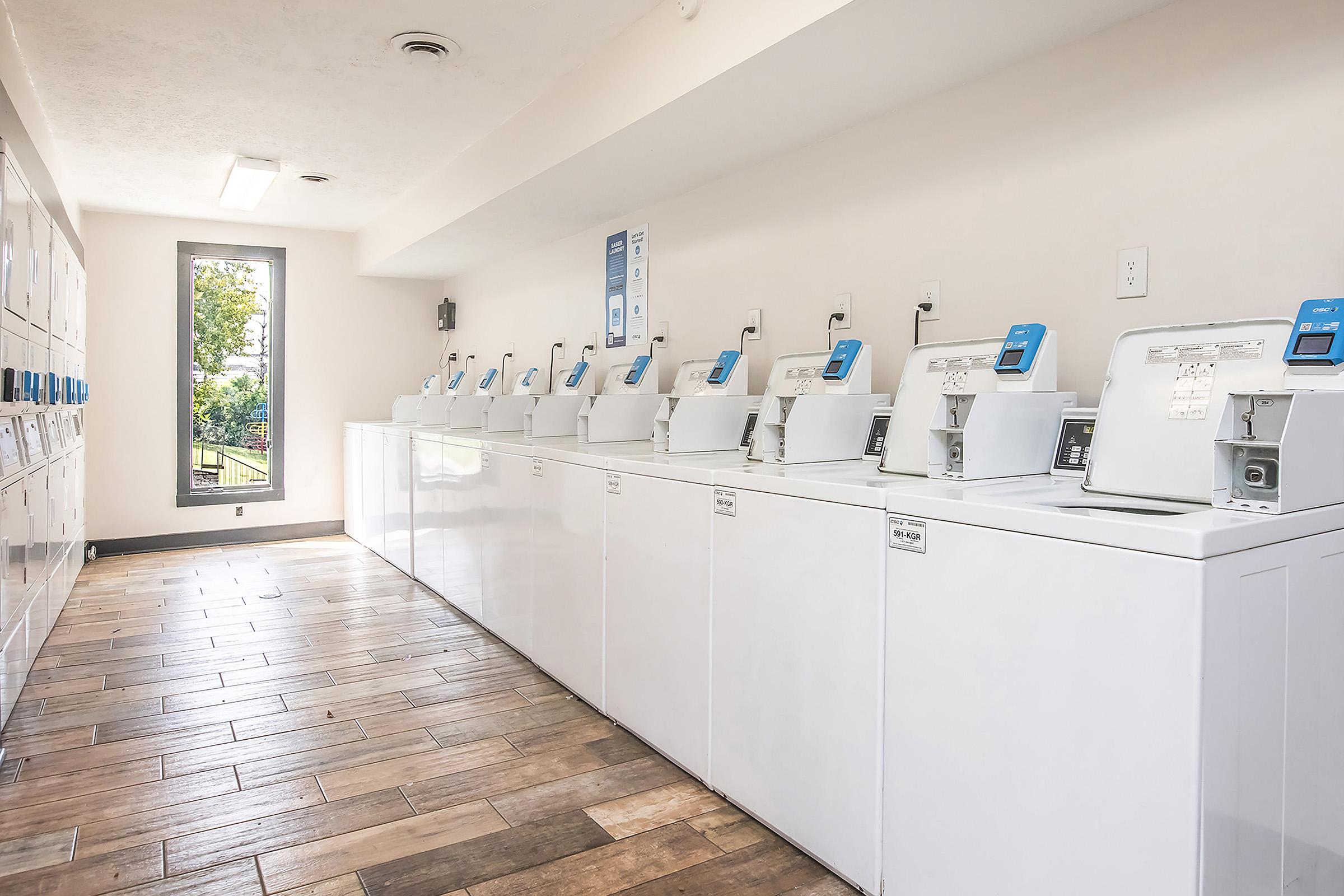 A clean and well-lit laundry room with multiple washing machines lined up against a wall. The floor is made of wooden planks, and there are windows allowing natural light to enter the space. The walls are painted in a neutral color, creating a modern and inviting atmosphere.