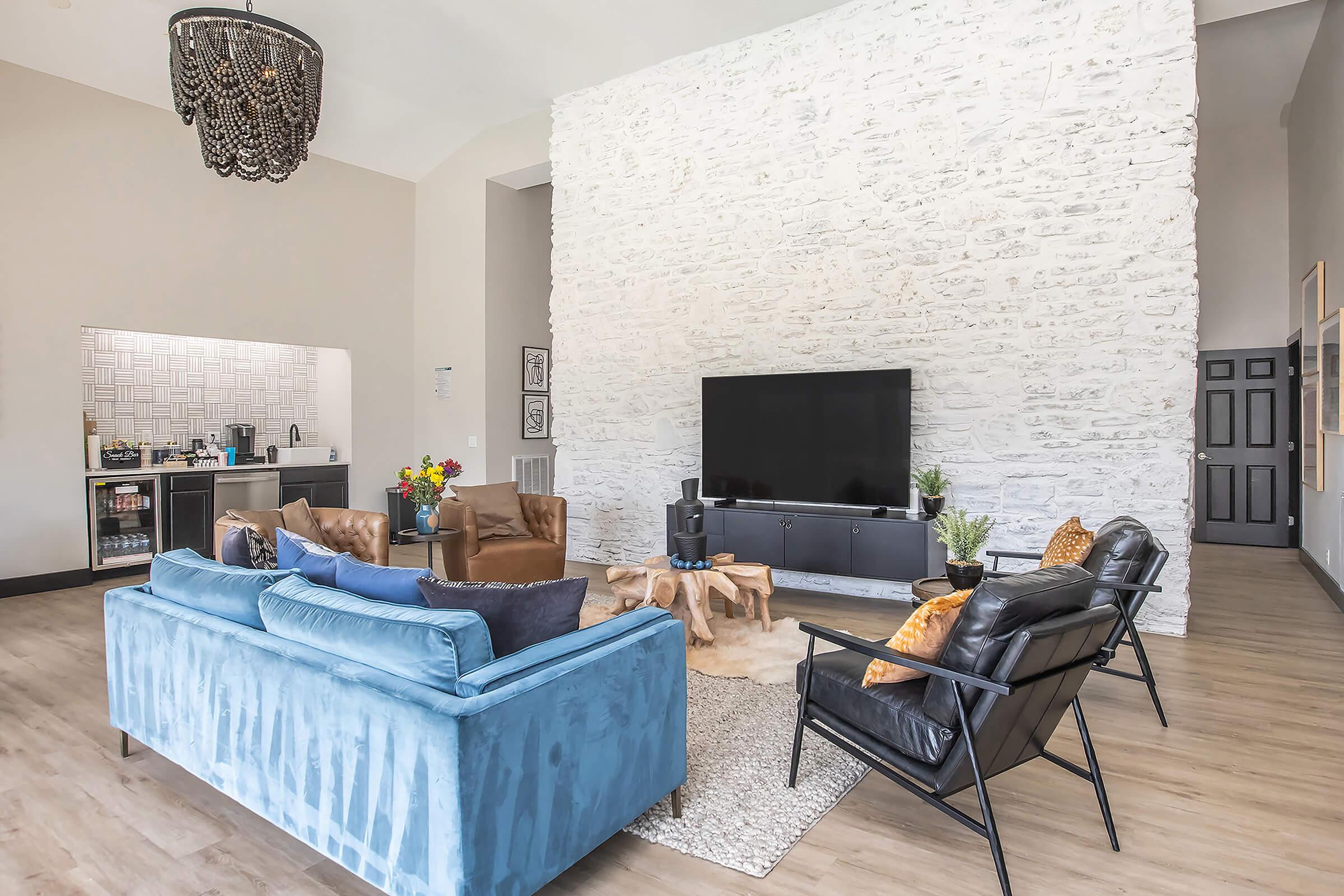 A modern living room featuring a large white stone wall, a flat-screen TV, and stylish furniture. There is a blue velvet sofa, two black armchairs, and a wooden coffee table. A cozy area rug lies beneath. The space is complemented by a chandelier and an open kitchen area in the background with a bar.