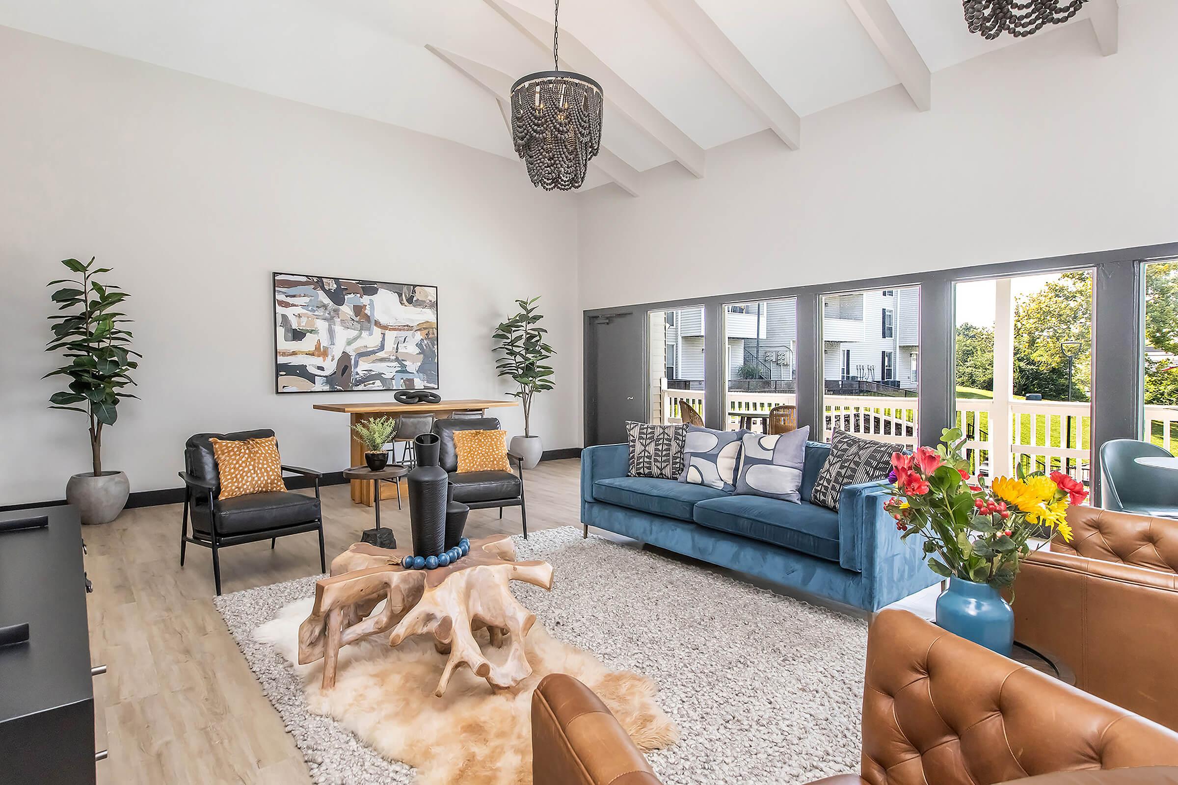 A modern living room featuring a blue sofa with decorative pillows, two leather armchairs, and a wooden coffee table with a unique design. There's a large area rug, plants in the corners, and a stylish chandelier overhead. Large windows offer a view of the outdoors, enhancing the bright and airy atmosphere.