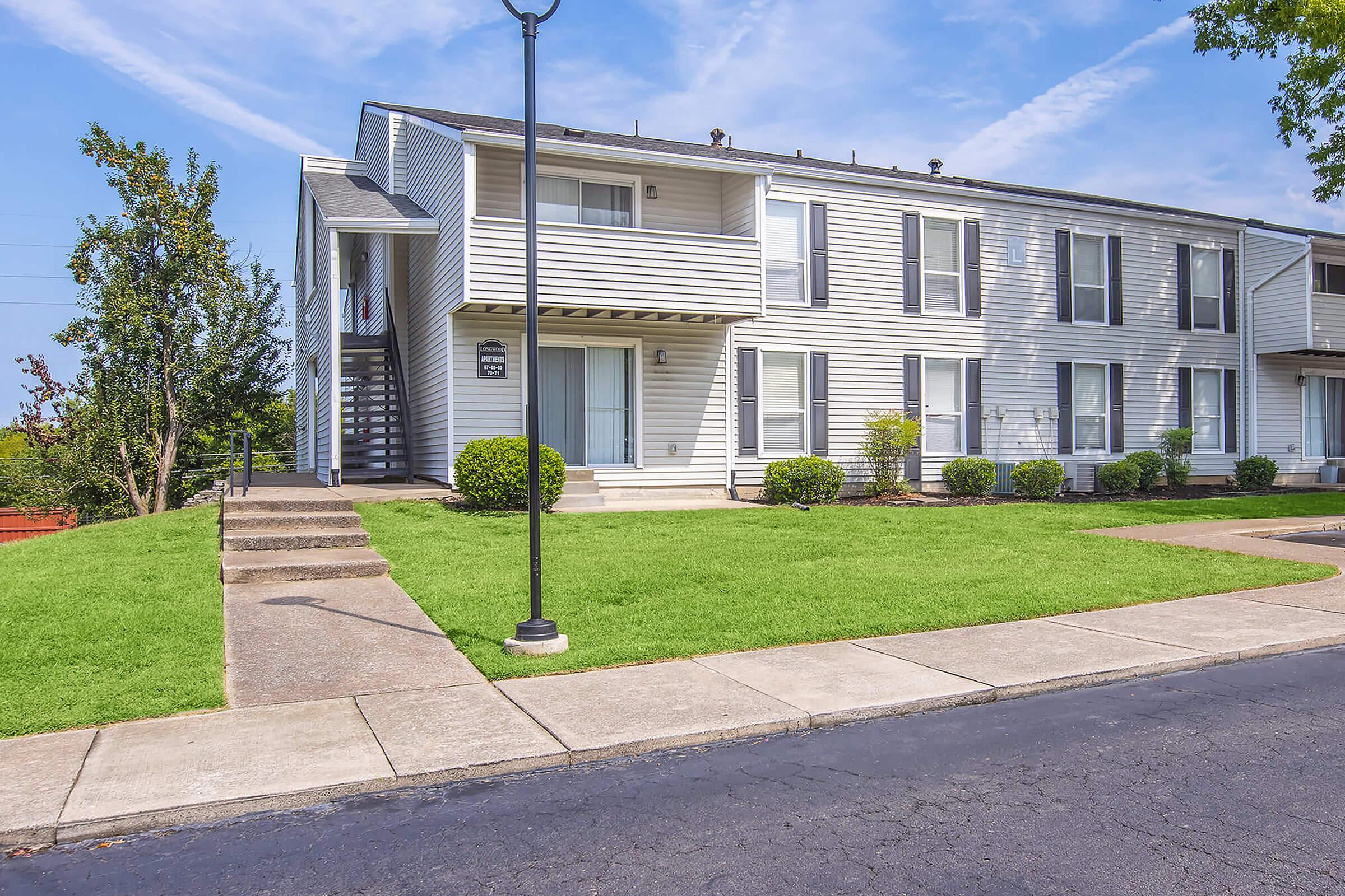 A two-story residential apartment building with a light-colored exterior. It features a set of stairs leading to the second floor, bordered by small hedges and green grass. A lamp post is near the sidewalk, which runs alongside the building and a patch of well-maintained lawn. Clear blue skies with a few clouds are visible.