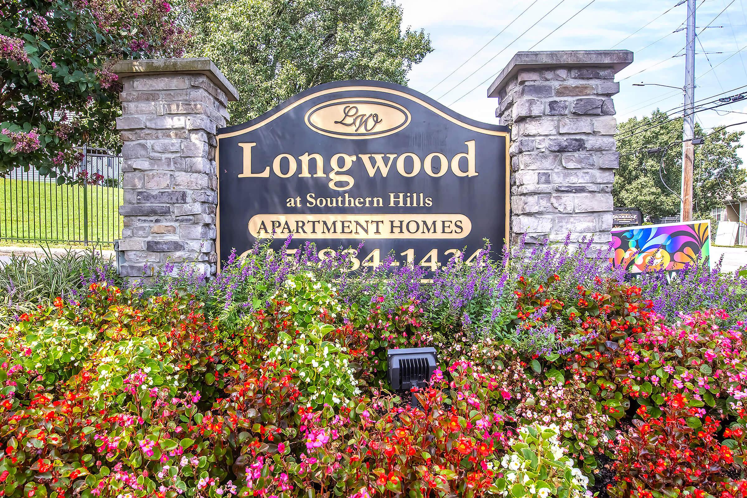 Sign for Longwood at Southern Hills Apartment Homes, featuring the name "Longwood" prominently displayed at the top. The sign is framed by decorative stone pillars and surrounded by vibrant flower beds with colorful blooms. The contact number 918-934-1436 is included.