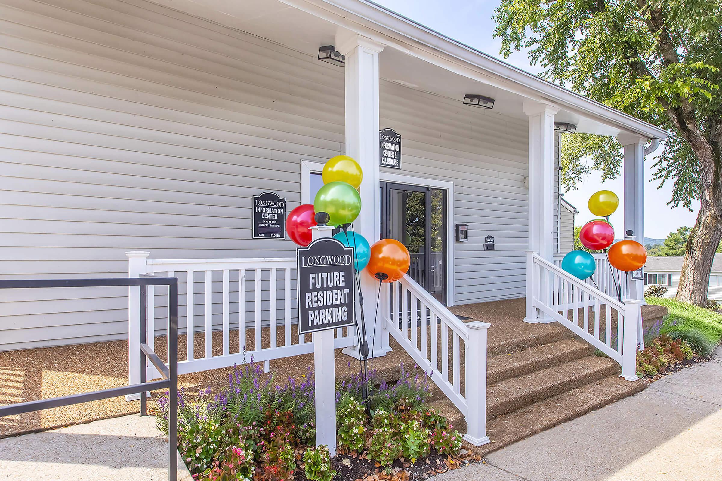 A white building with steps leading to the entrance, featuring a sign that reads "Future Resident Parking." Colorful balloons in orange, green, blue, and yellow are positioned near the entrance, adding a festive touch. The surrounding area includes flower beds with various plants.