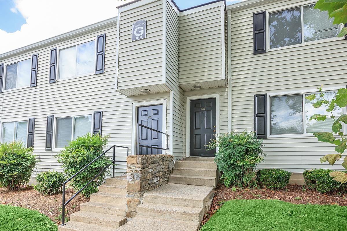 Exterior view of a multi-unit apartment building featuring a light-colored, horizontally striped siding. The entrance has a set of stairs leading up to a double door, flanked by black shutters on windows. The area is landscaped with green shrubs and grass, and the sky is partly cloudy.