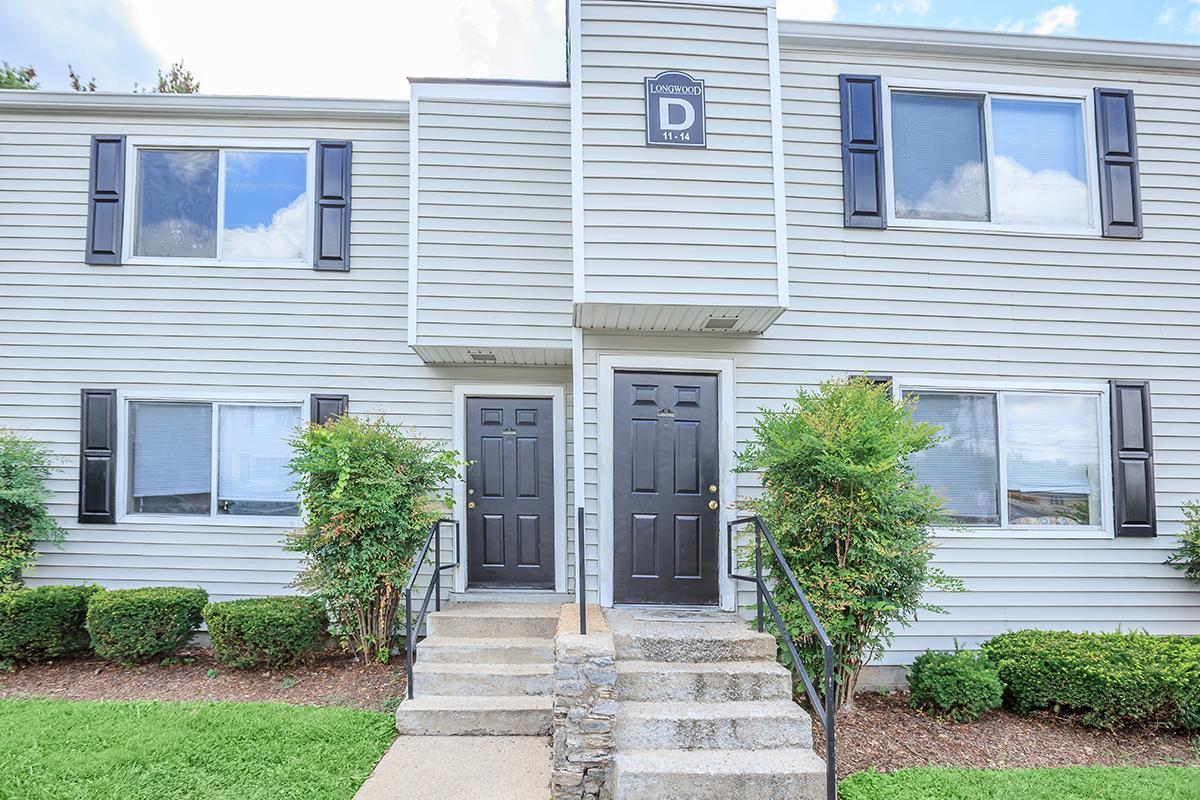 A two-story residential building with gray siding, featuring two front doors with black shutters. The entrance has concrete steps leading up to the doors. Lush green shrubbery and landscaping surround the entrance, with a clear blue sky visible in the background.