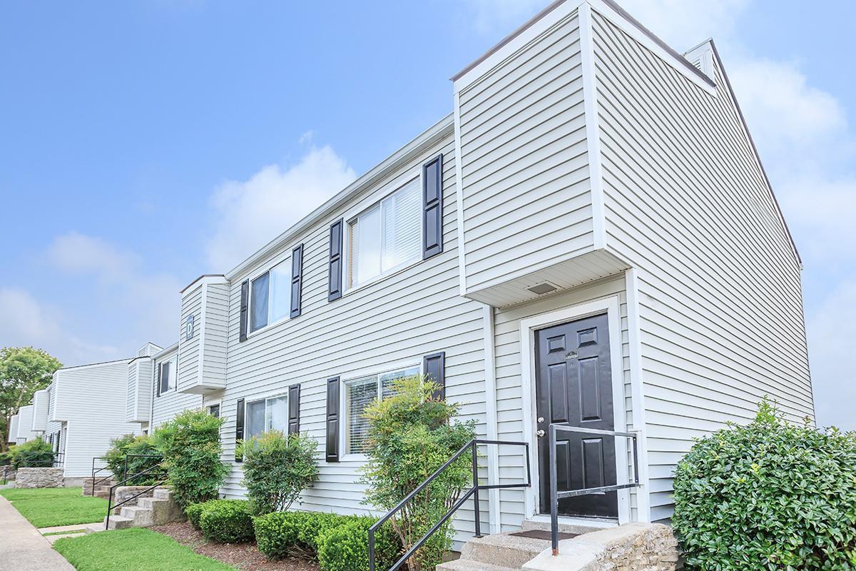 A modern two-story residential building with a light gray exterior, black doors, and windows, surrounded by small shrubs and a well-maintained path leading to the entrance. The sky is partly cloudy, and the building is part of a neighborhood with similar structures.