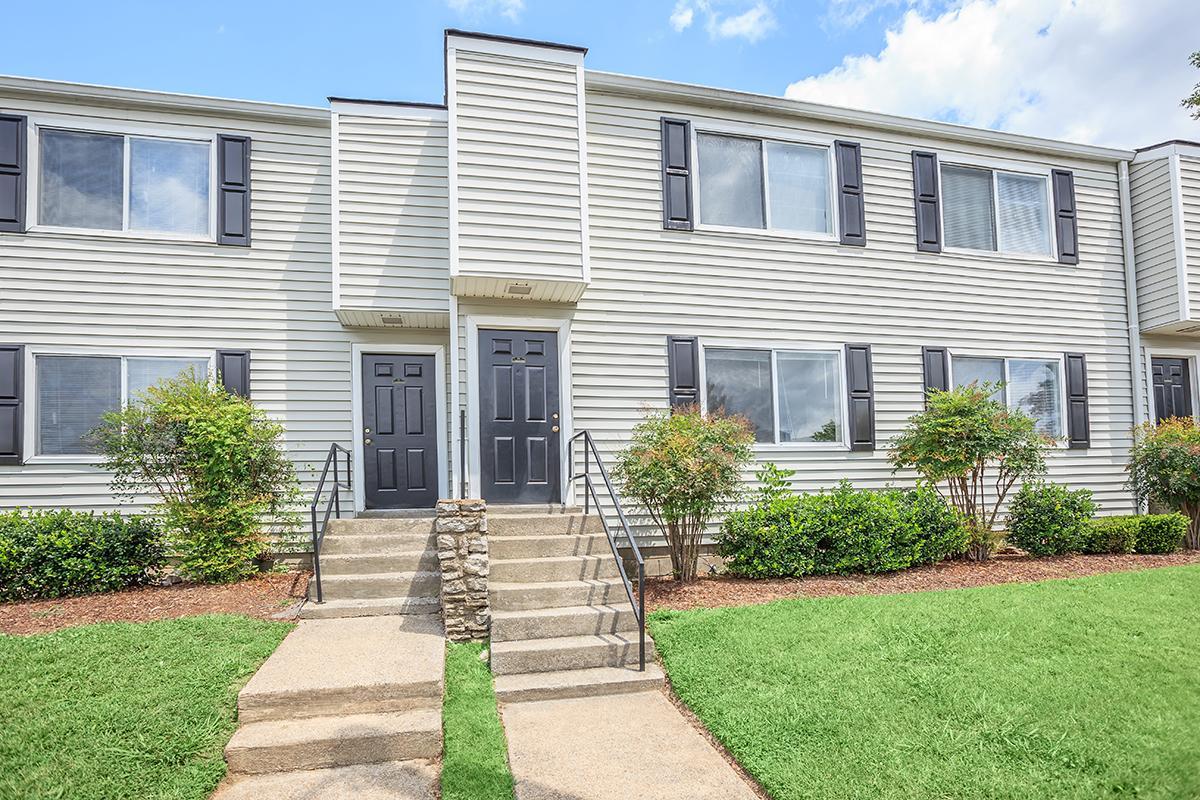 A two-story apartment building with light gray siding, featuring black shutters and doors. The facade includes several windows, and there are stone steps leading to the entrance. Lush green grass and well-maintained shrubs flank the pathway. The sky is partly cloudy.