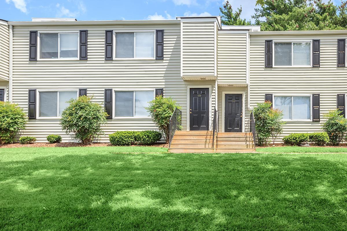A two-story apartment building with light-colored siding, featuring black doors and white-framed windows. Green grass and small shrubs surround the entrance, which has wooden steps leading up to the doors. The exterior reflects a well-maintained residential area with a clear blue sky in the background.