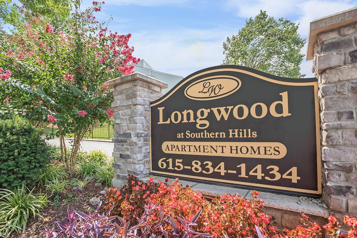 Sign for Longwood at Southern Hills Apartment Homes, featuring the name "Longwood" in elegant script, phone number 615-834-1434, and surrounded by vibrant flowerbeds and greenery. The setting includes a bright sky and trees in the background, creating a welcoming atmosphere.