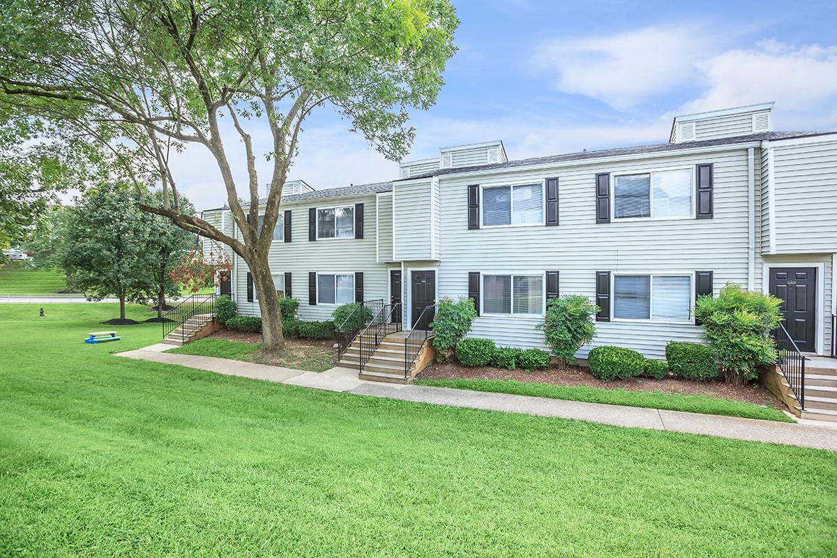 A low-rise apartment building with a light-colored exterior, featuring multiple units, black doors, and large windows. The surrounding landscape includes well-maintained grassy areas, a few trees, and decorative shrubs. A blue object is visible in the distance on the lawn.