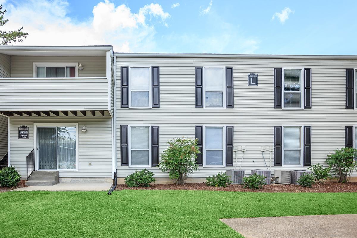 Two-story apartment building facade with multiple windows featuring black shutters. A covered porch area is on the left, leading to an entryway. The lawn is well-maintained with small shrubs, and the sky above is partly cloudy.