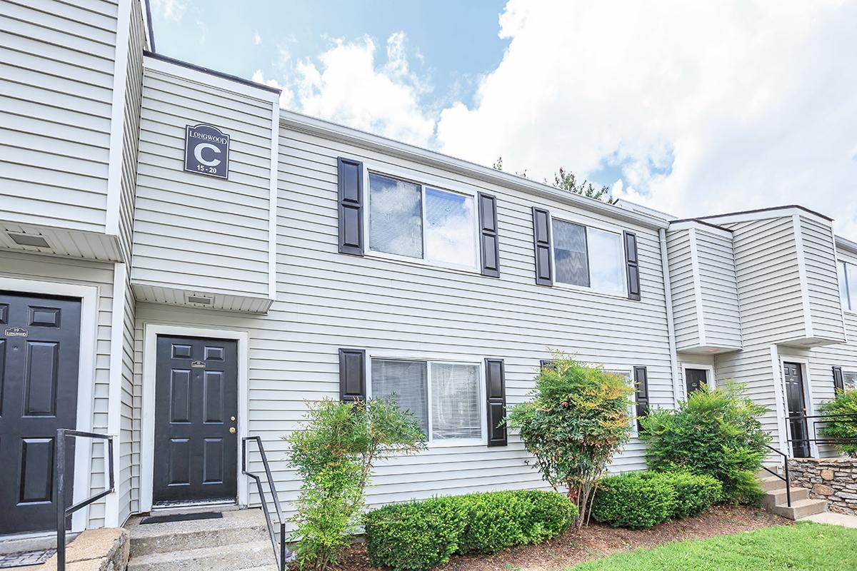 Exterior view of a two-story apartment building featuring light gray siding, black shutters, and a black front door. The building has a sign labeled "C" near the entrance. Lush green shrubs and small bushes are planted in front, with a clear blue sky and fluffy clouds above.