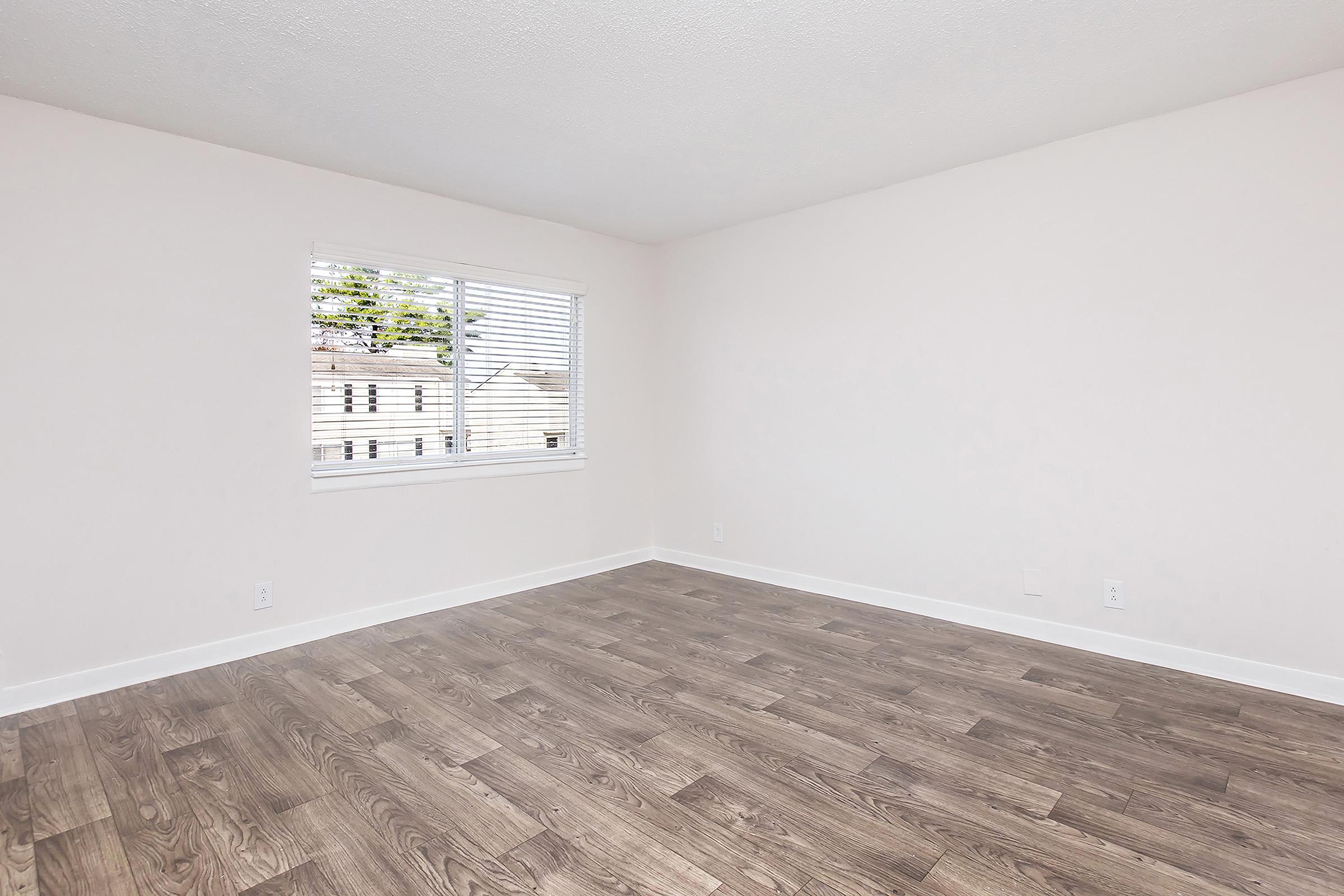 A bright, empty room with light-colored walls and a large window featuring white blinds. The flooring is a warm wood tone, creating a spacious and inviting atmosphere.