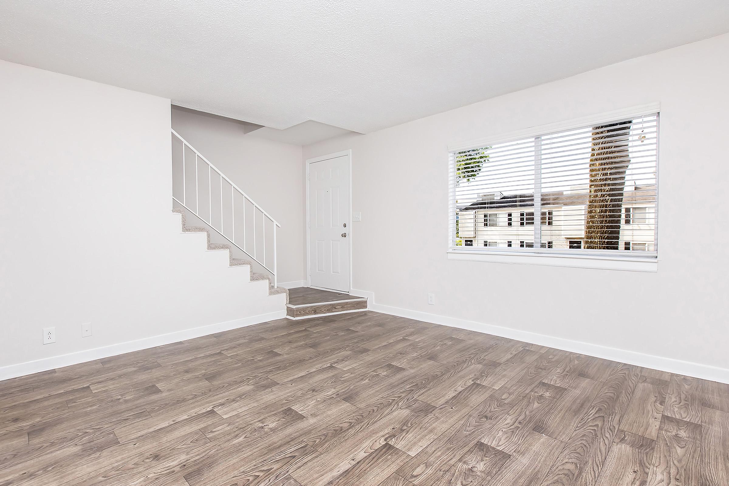 A spacious, minimalist living room with light-colored walls and hardwood flooring. There is a staircase in the corner leading to an upper level, and a large window with blinds allows natural light in, revealing a view of the outdoors. The room is empty, showcasing its potential for furnishing.
