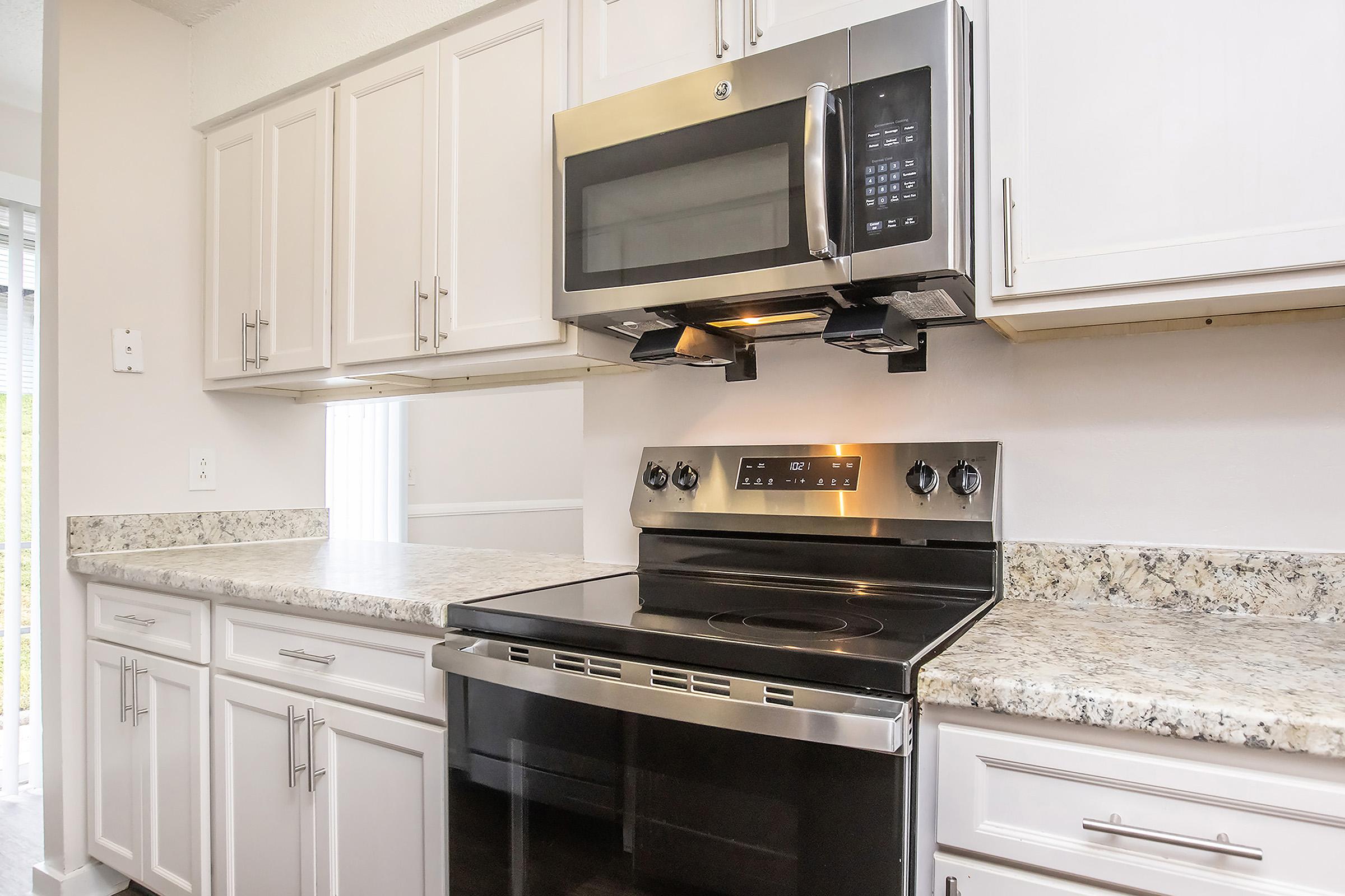 A modern kitchen featuring white cabinetry, a stainless steel microwave mounted above a black stovetop range, and a granite countertop. Natural light is coming from a window, enhancing the bright and spacious atmosphere of the area.