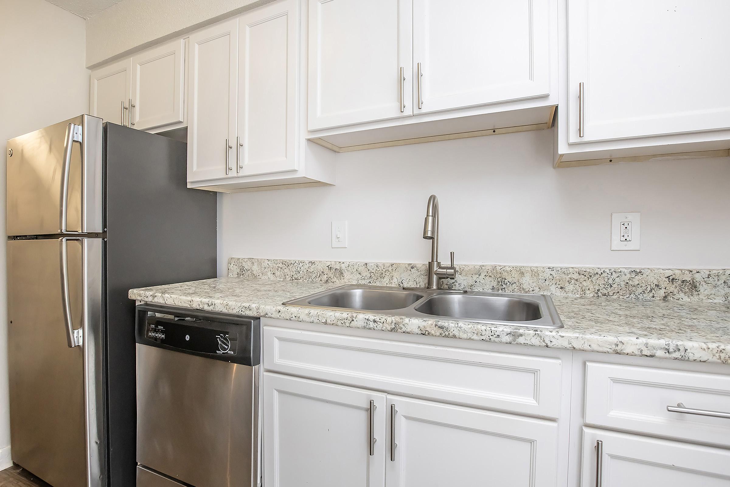 A modern kitchen featuring white cabinetry, a stainless steel refrigerator, and a dishwasher. The countertop is made of gray and white speckled granite, with a double sink and a stylish faucet. The overall design is clean and contemporary, creating a bright and functional cooking space.