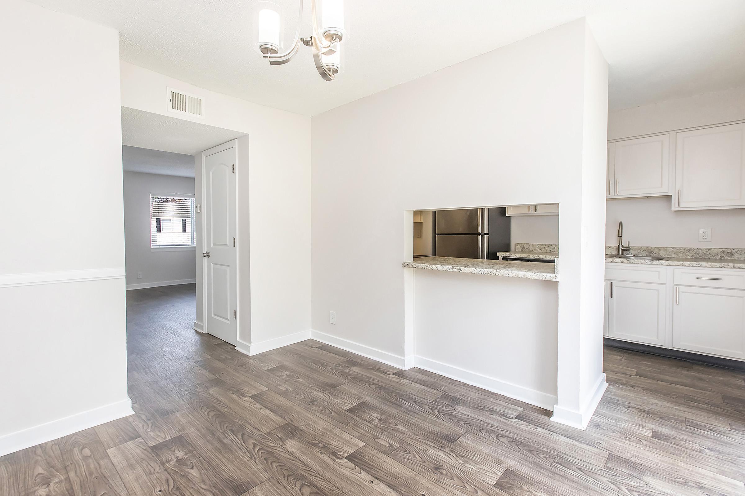 A bright, modern kitchen area with light wood flooring, featuring a granite countertop and a bar opening to a living space. The walls are painted white, enhancing the airy feel. A doorway leads to another room, and there are cabinets and appliances visible in the kitchen. A chandelier hangs from the ceiling.