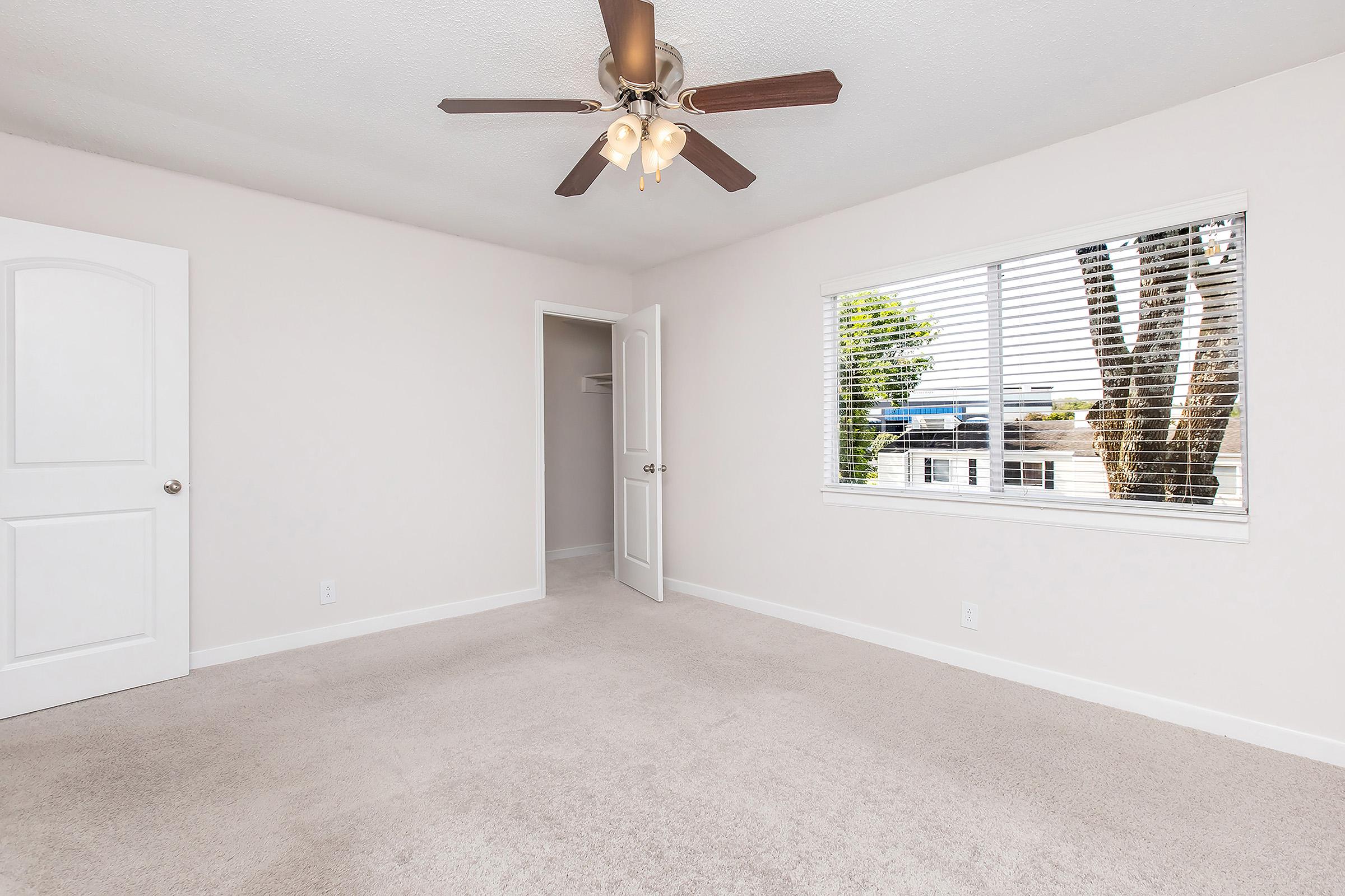 A well-lit, empty bedroom with light beige carpet and soft gray walls. It features a ceiling fan with light fixtures and a window with blinds, overlooking greenery outside. A partially open door leads to a closet space. The room has a spacious and clean appearance, ideal for personalization.