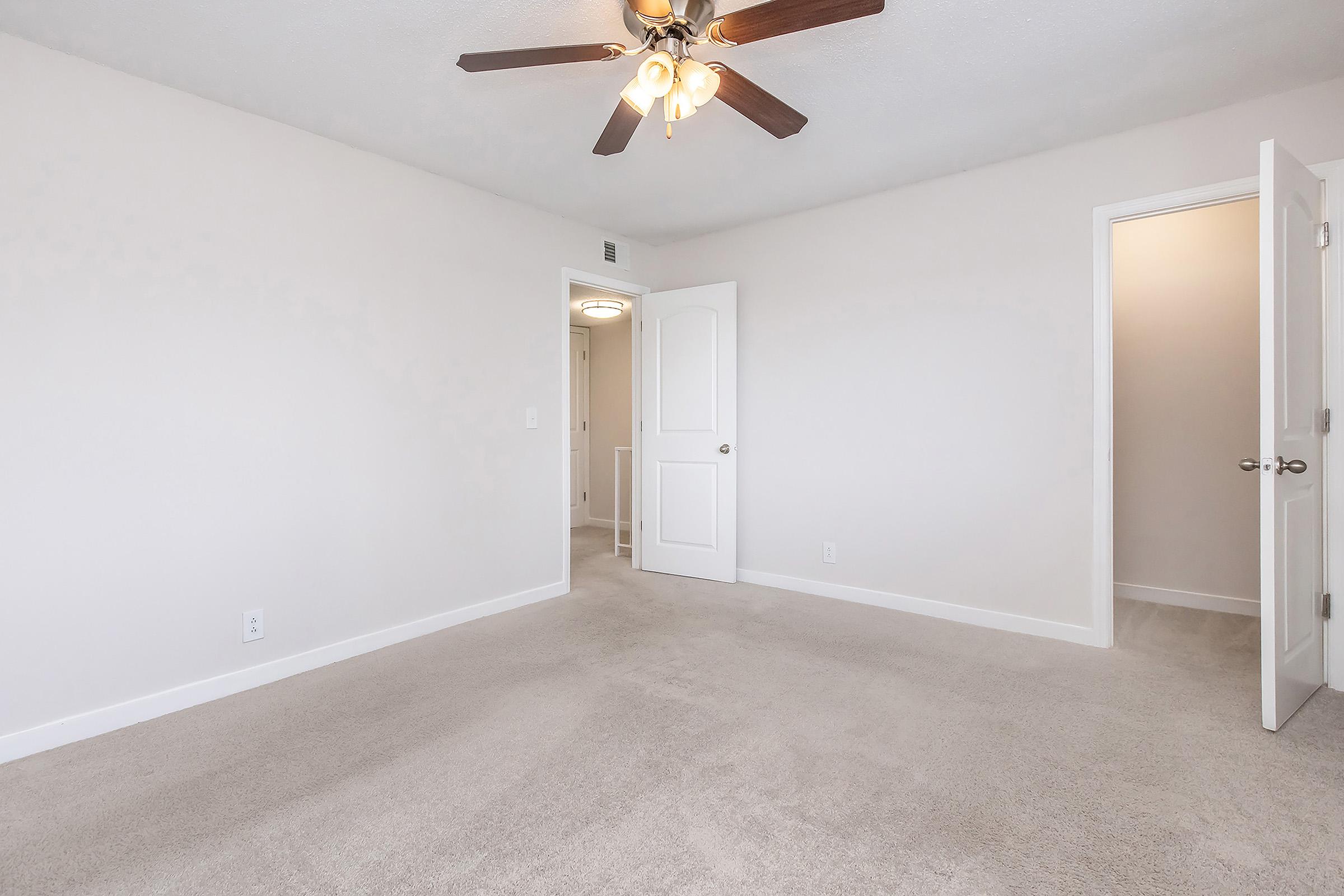 Empty room with light-colored walls and a light brown carpet. A ceiling fan with four lights is mounted above. Two doors are visible; one slightly ajar leading to another room and the other closed. The space has a bright and airy feel, suitable for various uses such as a bedroom or office.
