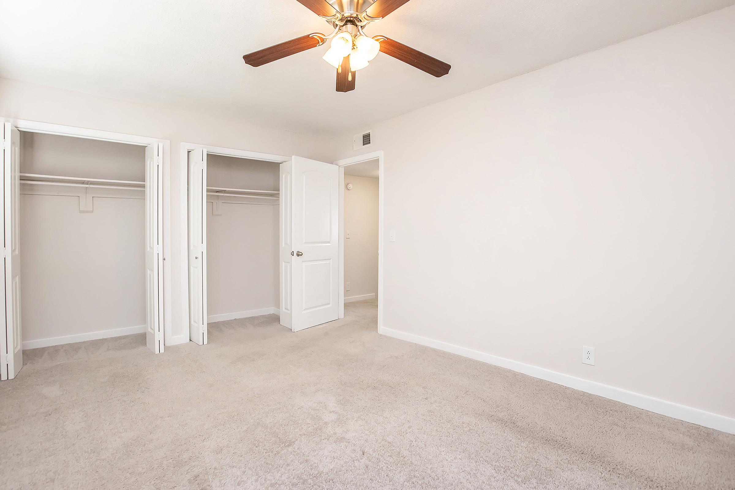 Empty room featuring light-colored walls and carpet, with a ceiling fan. Two open closet doors on the left side reveal spacious closets. The room has neutral decor and natural light coming in, creating a bright and airy atmosphere.