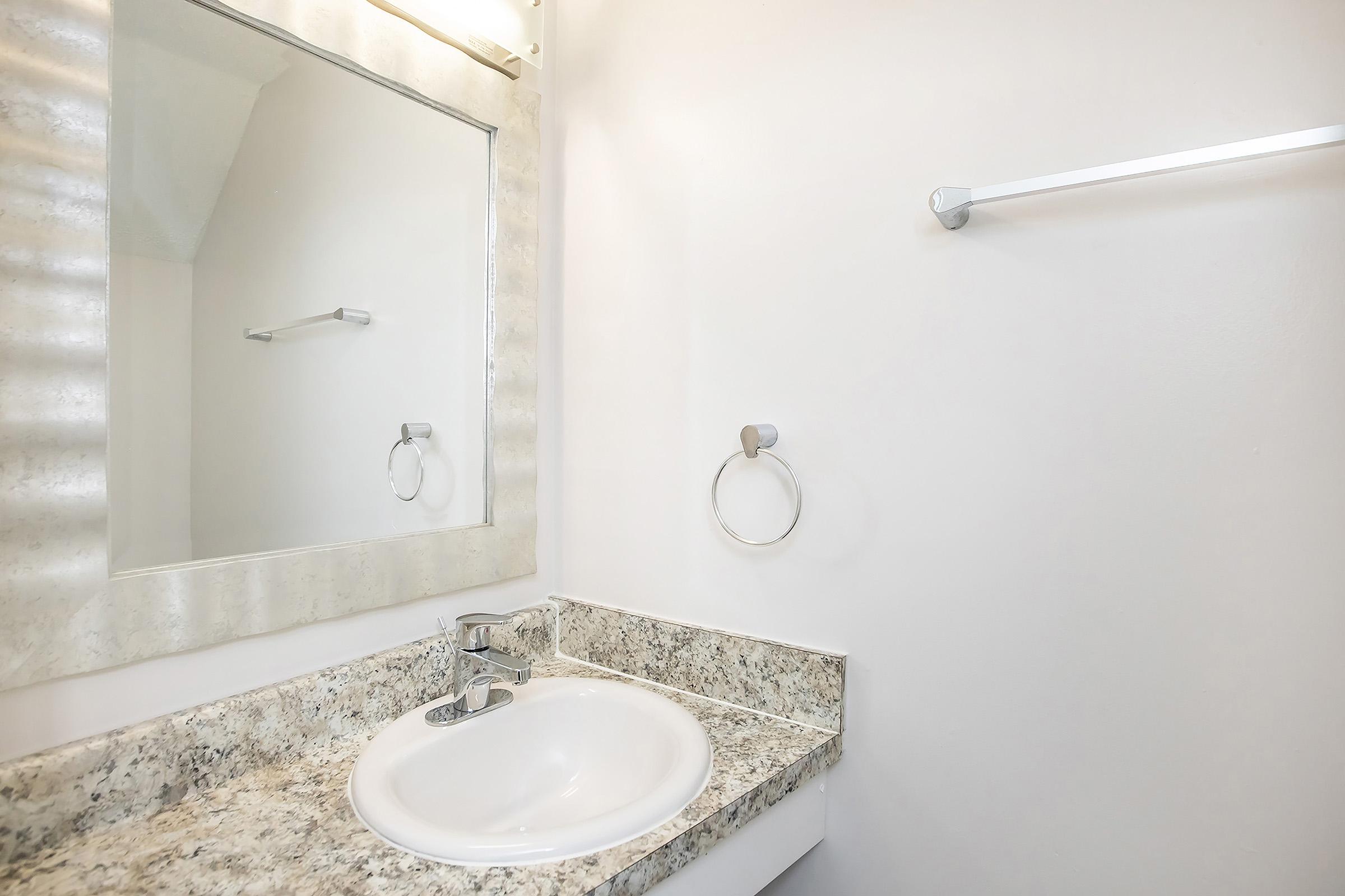 A modern, minimalist bathroom featuring a white sink with a polished chrome faucet, a round mirror, and a towel bar. The countertop is made of speckled granite, and the walls are painted in a light neutral color to enhance the space's brightness.