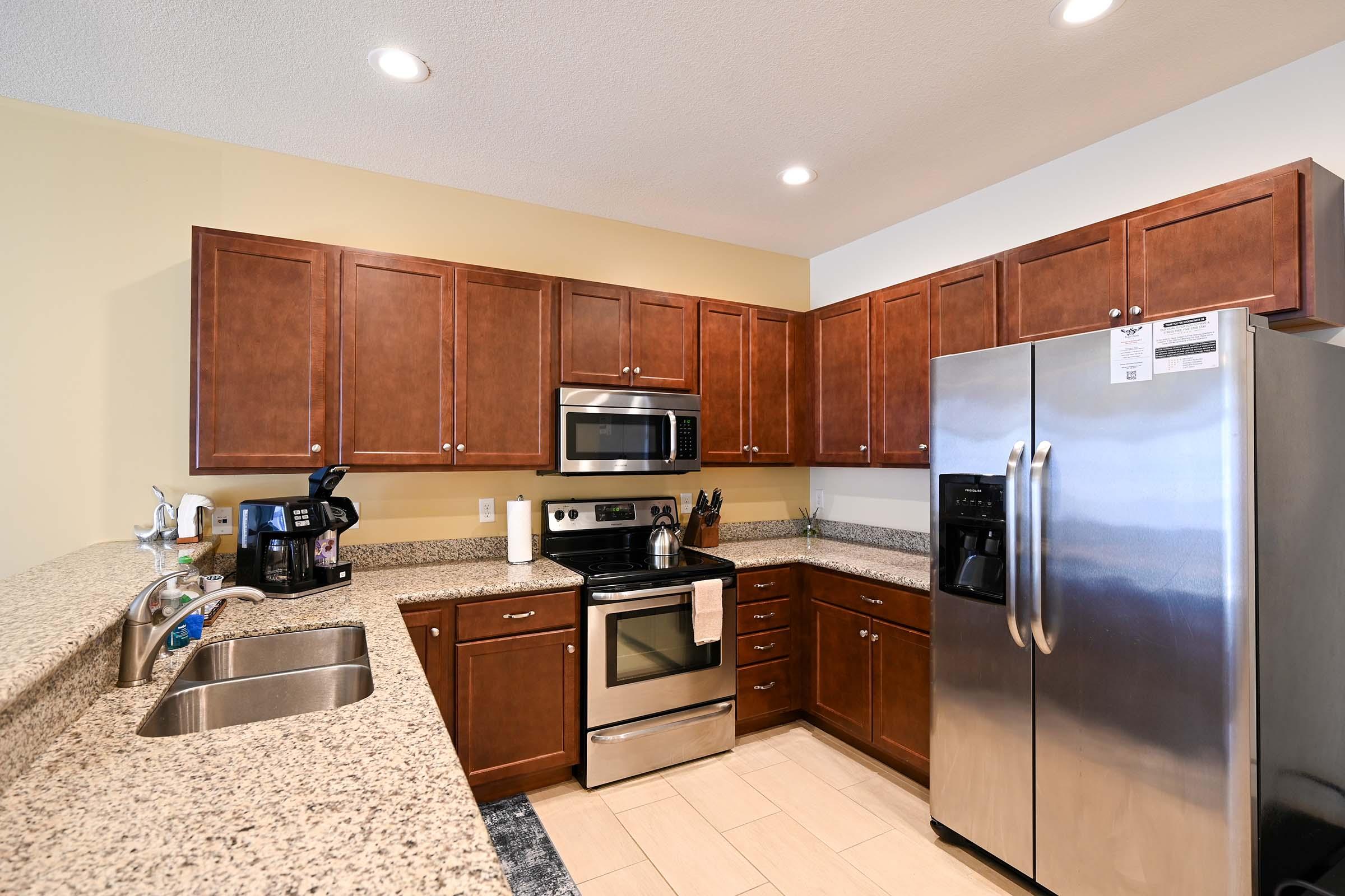 Modern kitchen featuring dark wood cabinets, granite countertops, stainless steel appliances including a refrigerator, oven, microwave, and a coffee maker, with a double sink and light-colored walls. The layout is functional, showcasing an organized and well-equipped cooking space.