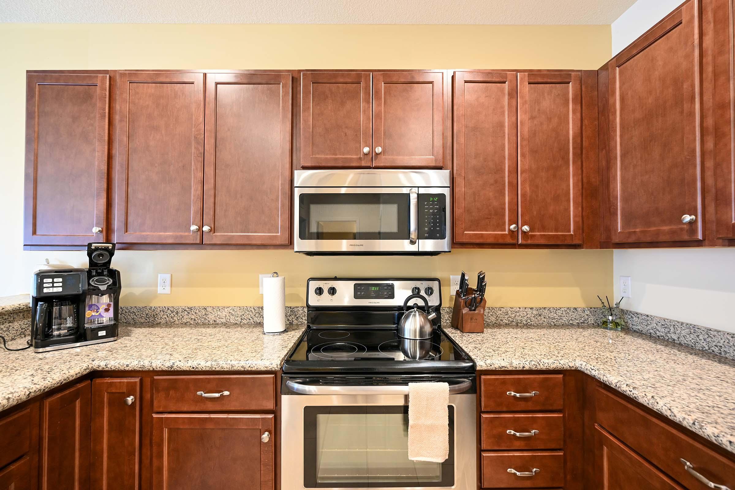 A modern kitchen featuring wooden cabinets, a stainless steel stove and microwave, and a granite countertop. A coffee maker is positioned alongside a utensil holder, and a kettle sits on the stove. The overall decor is clean and contemporary, with light yellow walls enhancing the warm wood tones.