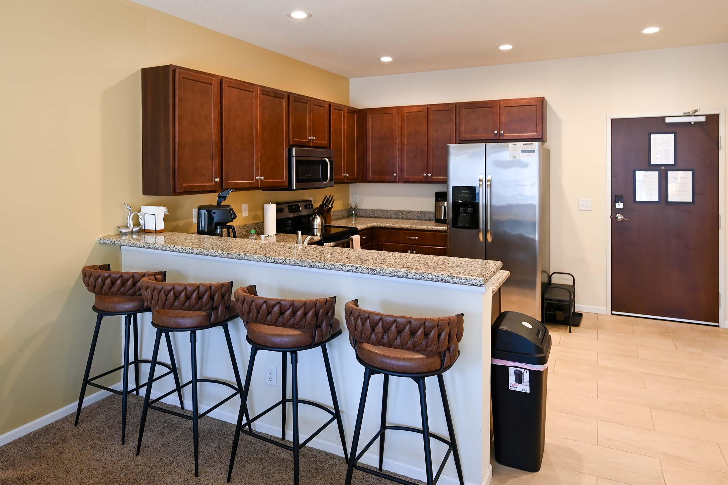 Modern kitchen featuring dark wood cabinets, stainless steel appliances, and a granite countertop. There are five high stools with brown leather upholstery at the breakfast bar. In the background, a door leads to an exit, and a trash bin is visible near the kitchen area. The flooring is light-colored tile.