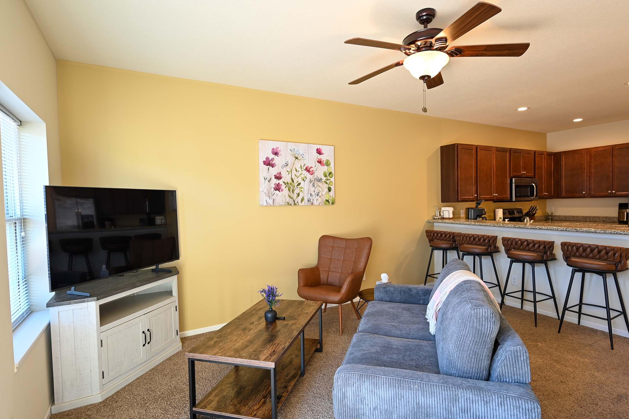 A cozy living room featuring a gray sofa and a brown armchair. There's a wooden coffee table and a flat-screen TV on a stand. In the background, a kitchen bar with three padded stools is visible. A floral artwork hangs on the yellow wall, adding a touch of color to the space.