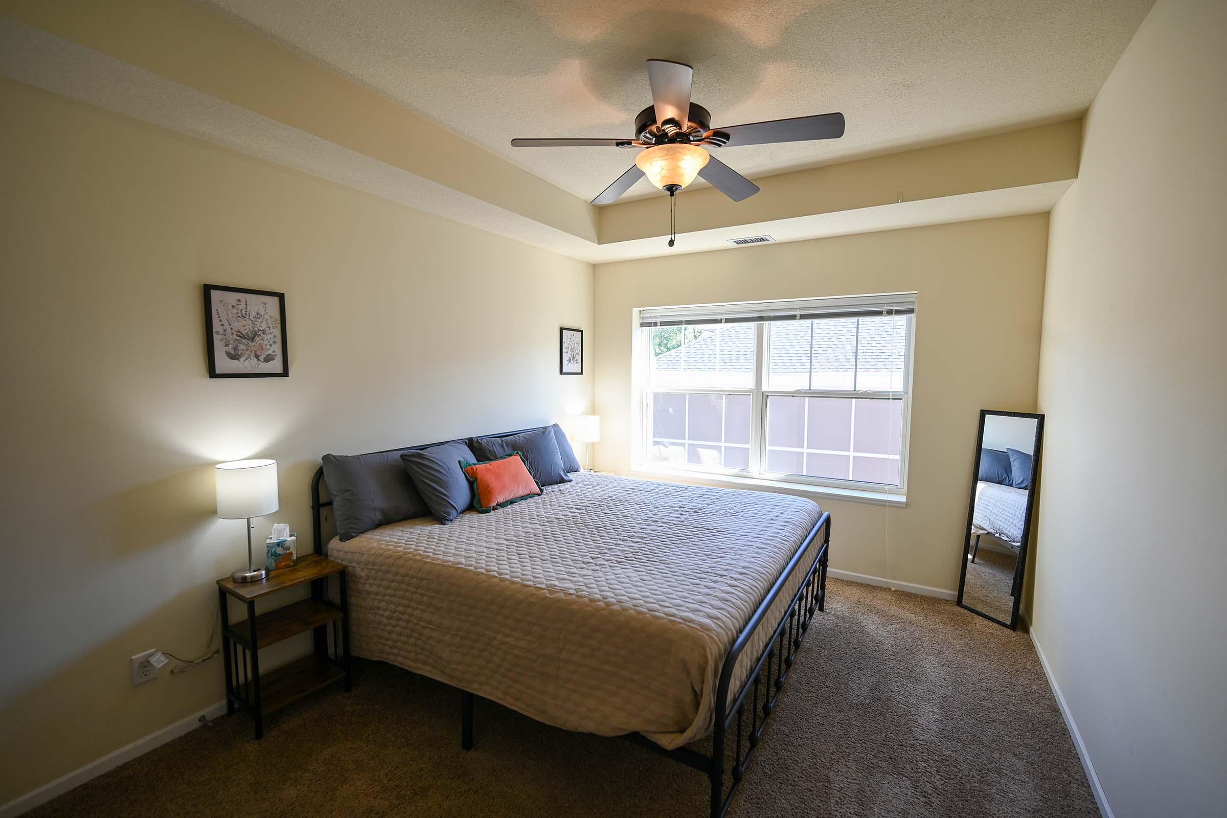 A cozy bedroom featuring a large bed with gray bedding and an orange accent pillow. There is a nightstand with a lamp beside the bed, a mirror on the floor, and a window allowing natural light to fill the room. The walls are painted in a neutral color, enhancing the inviting atmosphere.