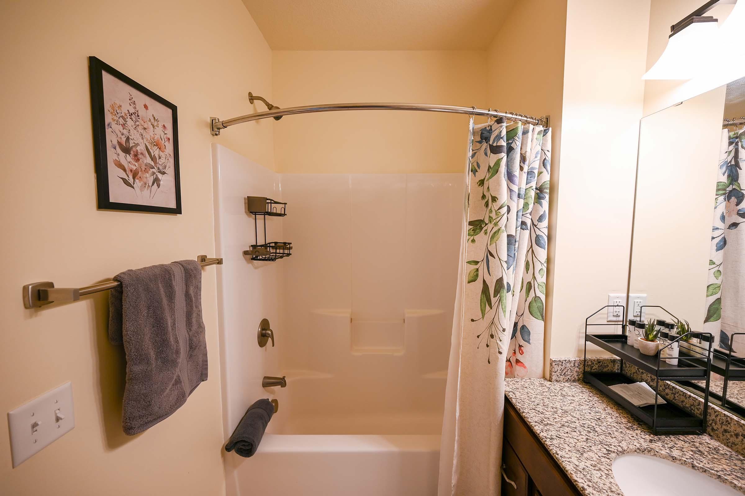 A clean bathroom featuring a shower with a curved curtain rod and a floral shower curtain. There are two wall-mounted shelves holding toiletries, a dark gray towel hanging on a rack, and a neatly organized vanity with a mirror. The decor includes a framed floral artwork.