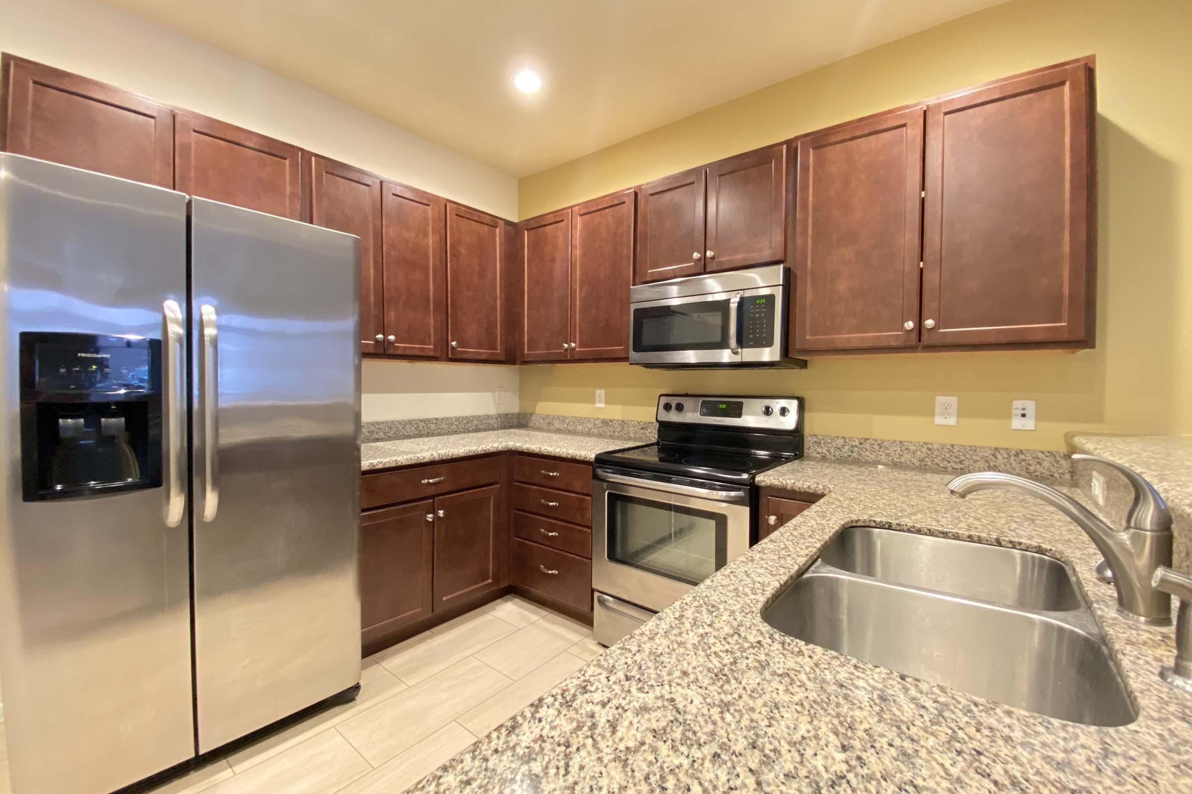 Modern kitchen featuring dark wood cabinets, stainless steel appliances including a refrigerator, oven, and microwave, and a granite countertop with a double sink. Soft yellow walls complement the warm tones of the cabinetry, creating a welcoming atmosphere. Bright overhead lighting enhances the space.