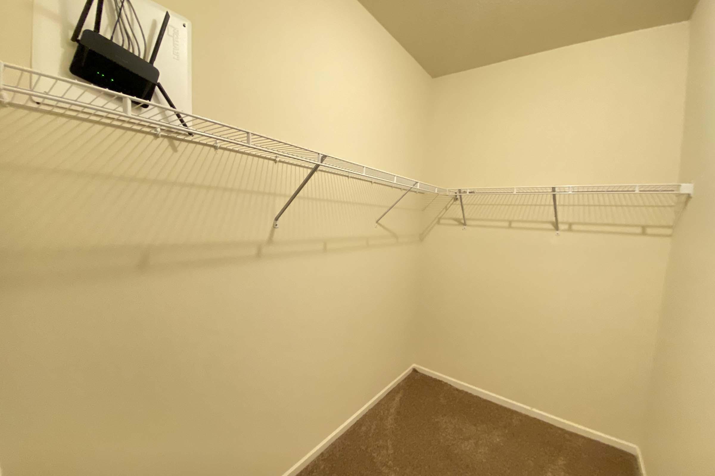 An empty closet with beige walls and a brown carpet. There is a wire shelving unit along one wall, with one shelf mounted higher than the others. A router is mounted on the wall above the shelves. The space is brightly lit and appears tidy.