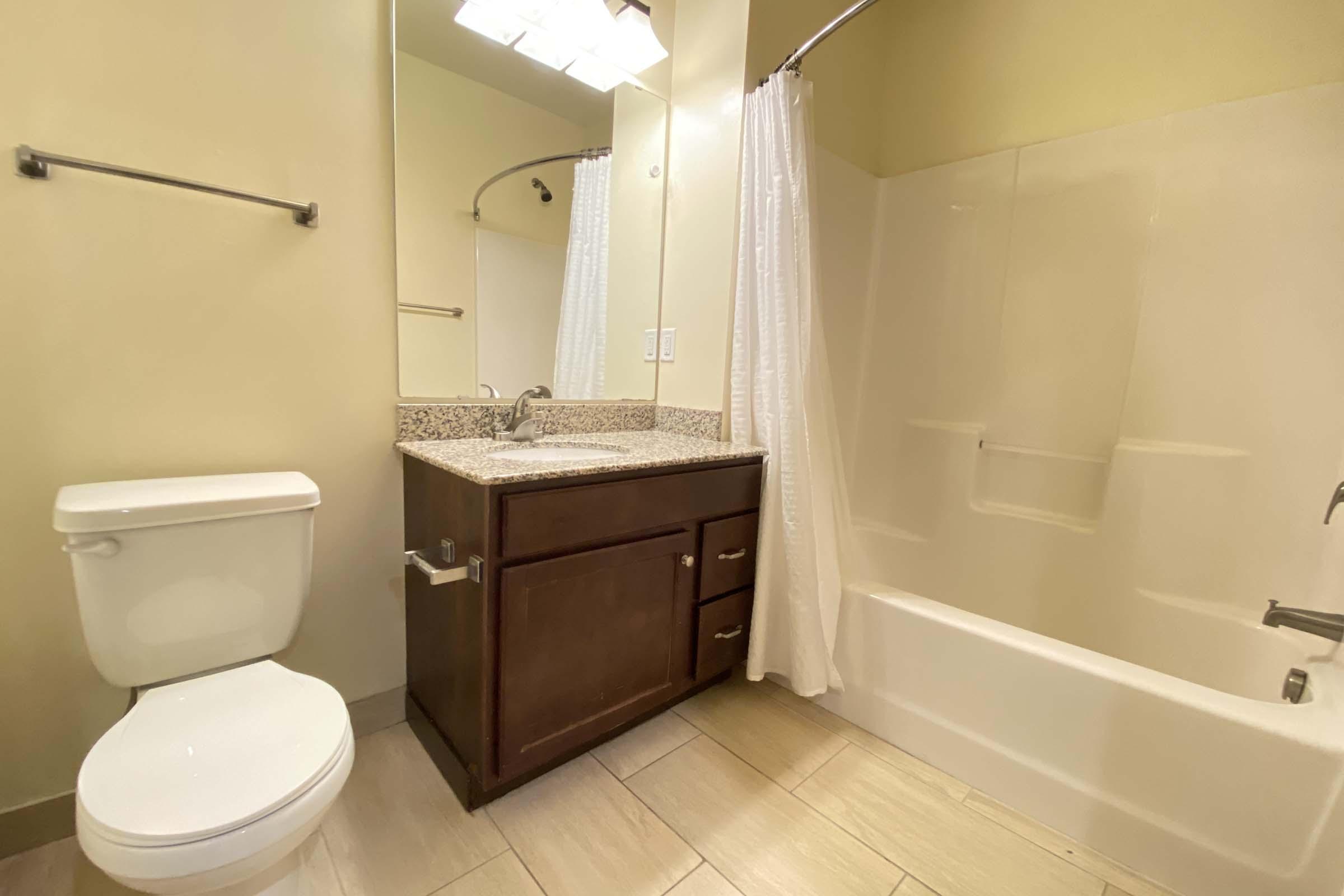 A clean and modern bathroom featuring a white toilet, a dark wood vanity with a sink, and a shower/tub combination with a white curtain. The walls are painted in a light yellow, and there are neutral-toned tiles on the floor. A mirror is above the vanity.