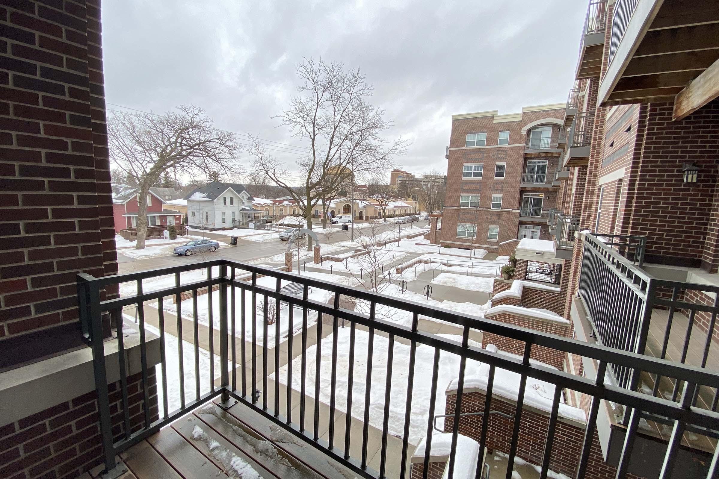 View from a balcony overlooking a snowy street. The scene features a mix of residential buildings, including houses and apartments, with snow-covered ground and bare trees. The sky is overcast, hinting at a cold day. A few parked cars are visible in the distance, and the overall mood is quiet and serene.